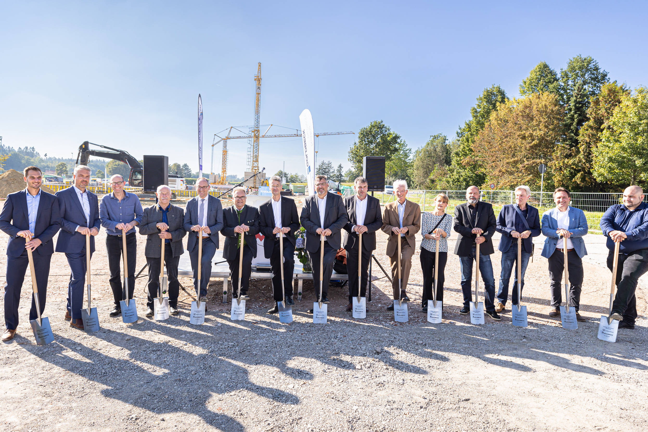 Über zehn Menschen stehen nebeneinander vor einer Baustelle, alle haben einen Spaten in der Hand, die Sonne scheint