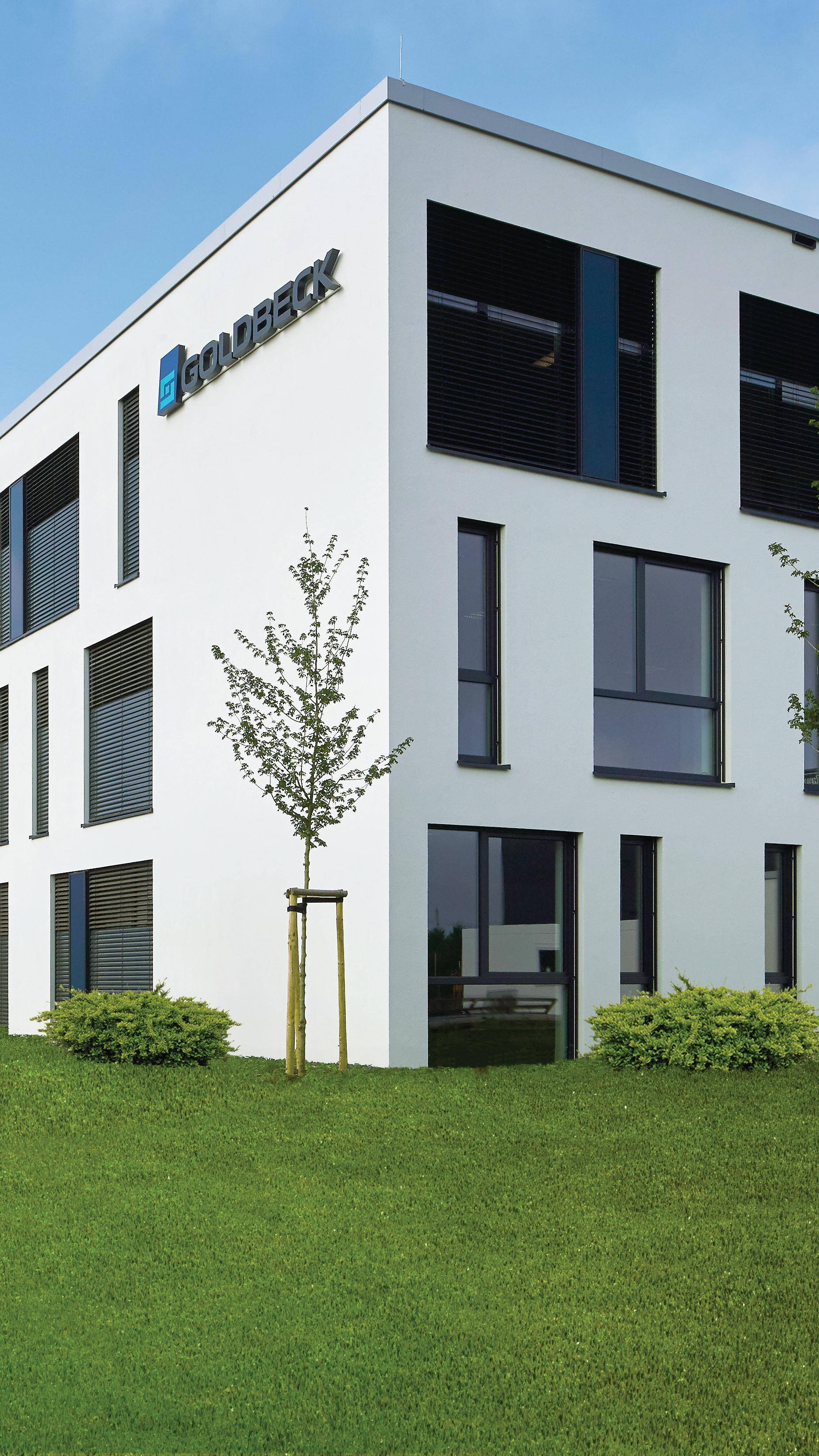 Modernes Bürogebäude mit weißer Fassade, großen Fenstern und kleiner Grünanlage im Vordergrund.