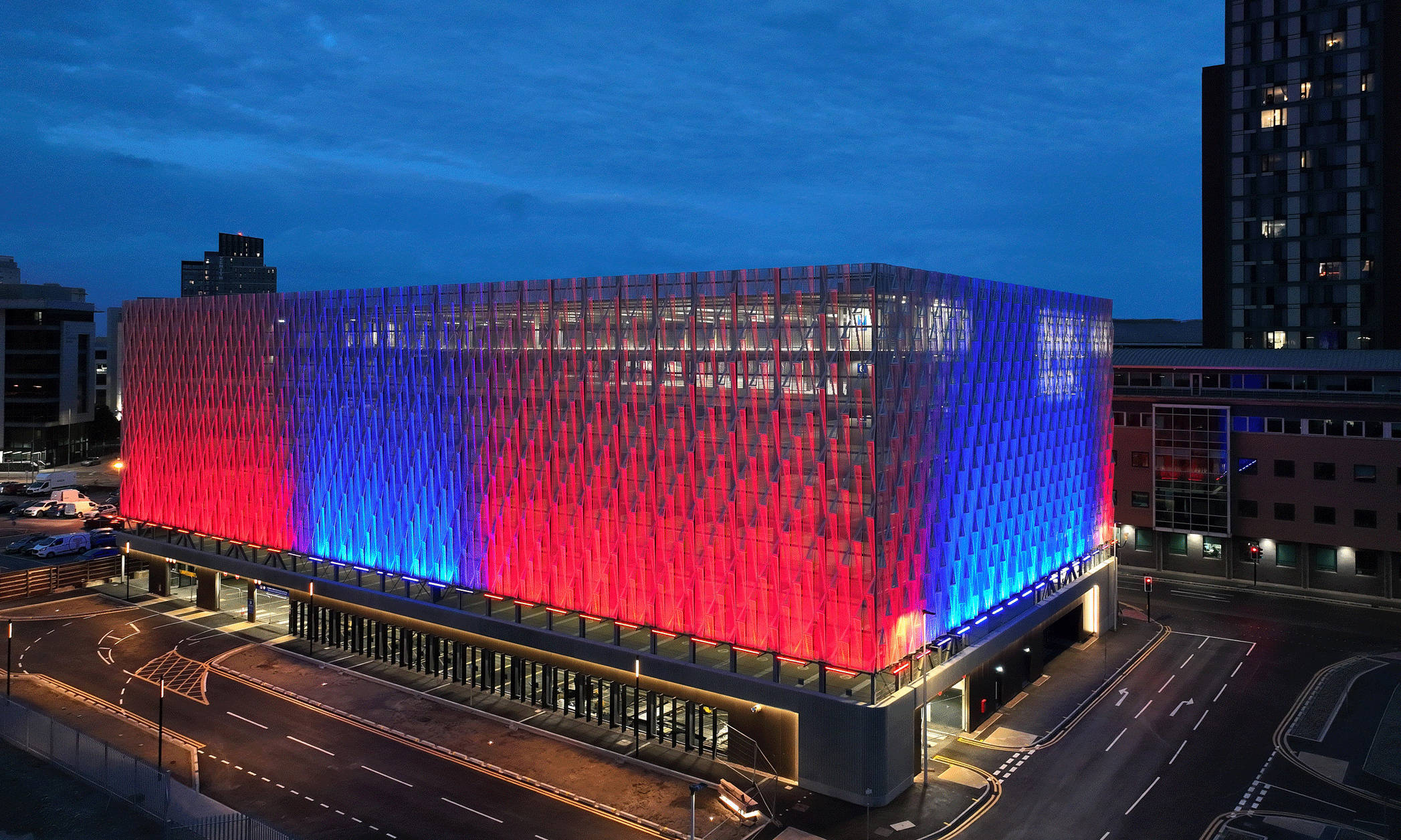 A modern building with vivid red and blue lighting at night, surrounded by streets and skyscrapers.