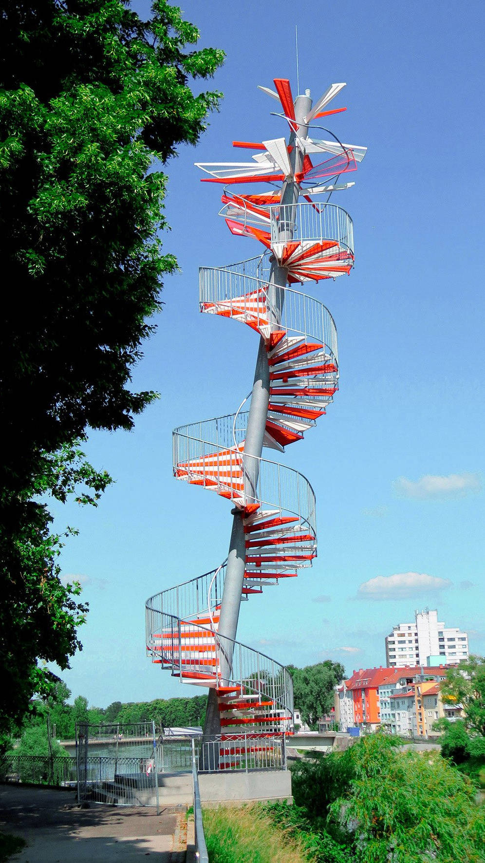 Spiralförmige Treppe mit buntem Handlauf vor blauem Himmel, umgeben von Grün und urbaner Architektur im Hintergrund.