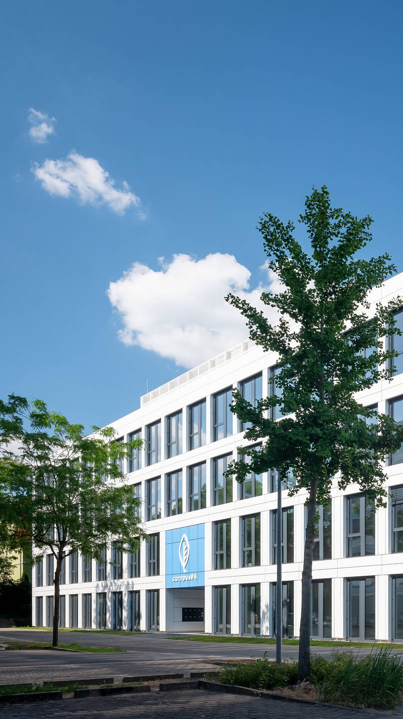 Modernes Bürogebäude mit Glasfassade vor blauem Himmel und einigen Bäumen im Vordergrund.