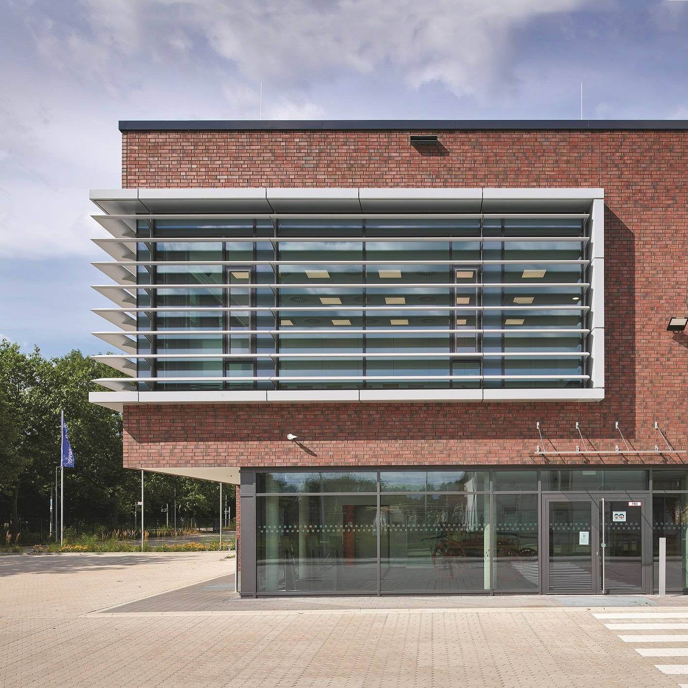 Modern brick façade of a building with large windows and horizontal slats, surrounded by trees.