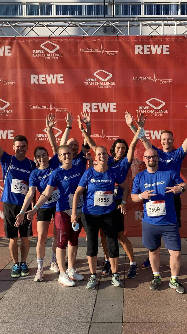 Eine Gruppe von acht Teilnehmern in blauen T-Shirts steht jubelnd vor einem roten Banner der REWE Team Challenge Dresden.