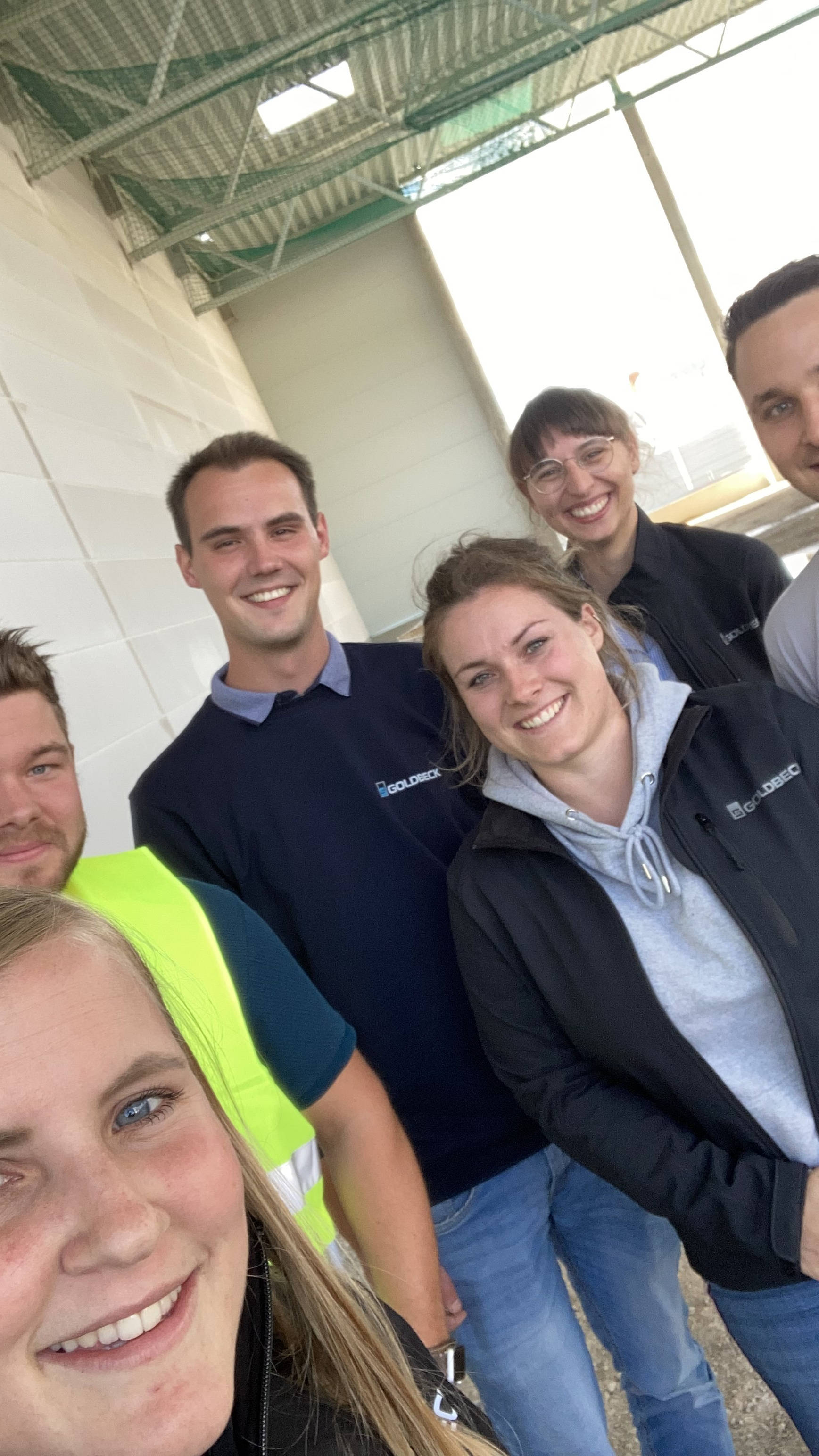 Gruppe von sechs lächelnden Personen in Arbeitskleidung vor einem Gebäude.