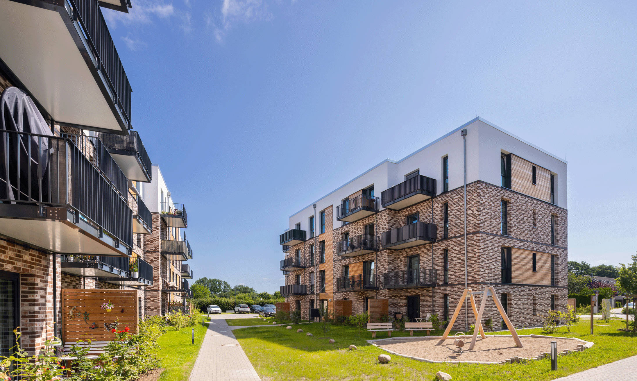 Modern residential complex with multi-storey buildings, gardens and children's playground in sunny weather.