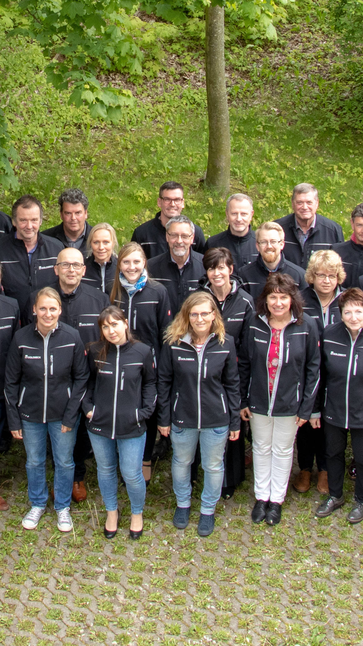 Gruppe von Menschen in identischen schwarzen Jacken steht draußen auf einem gepflasterten Weg, umgeben von Bäumen.