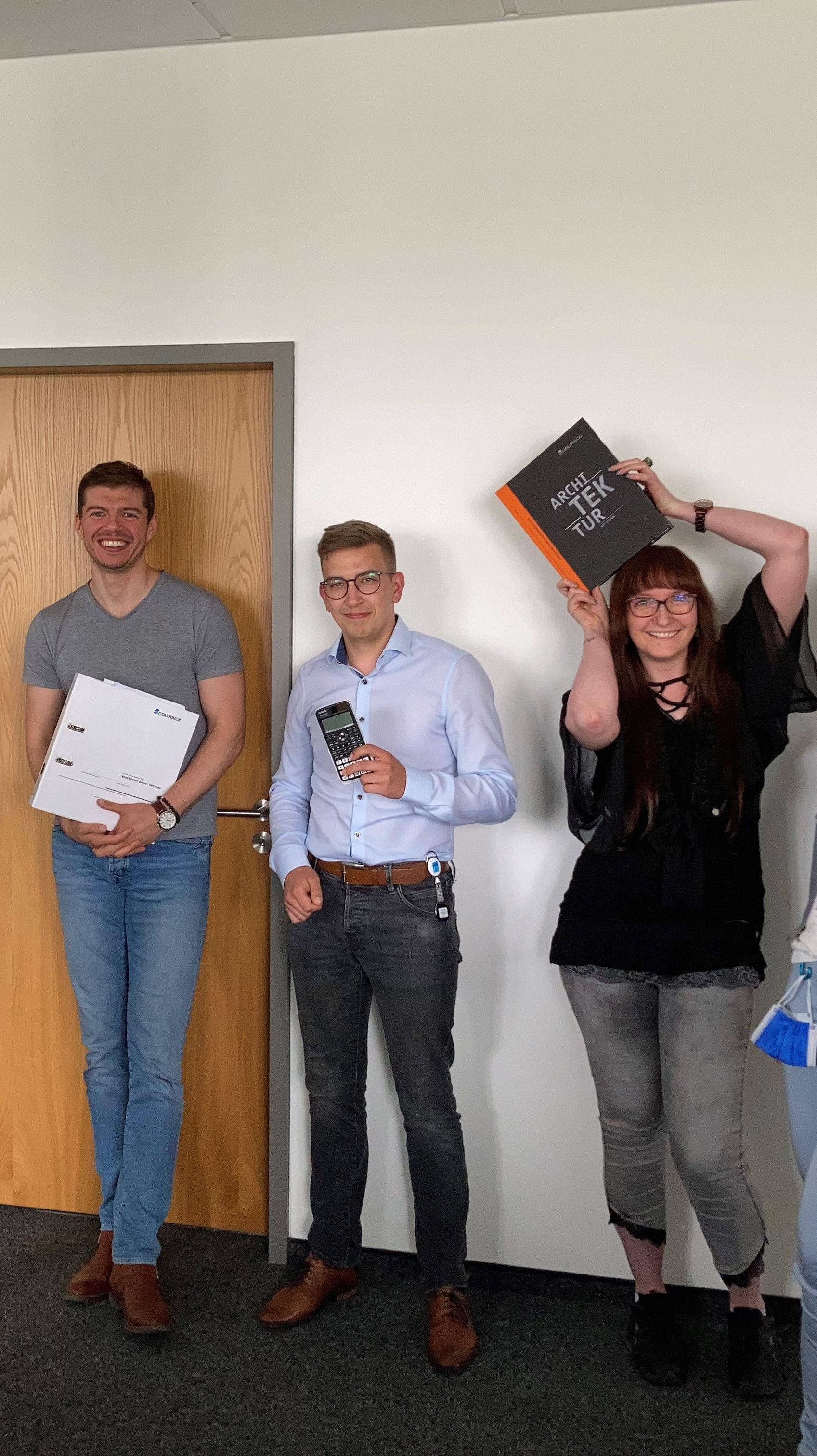 Eine Gruppe von sechs Personen steht lächelnd in einem Büroflur, einige halten Bücher und Farbmuster.