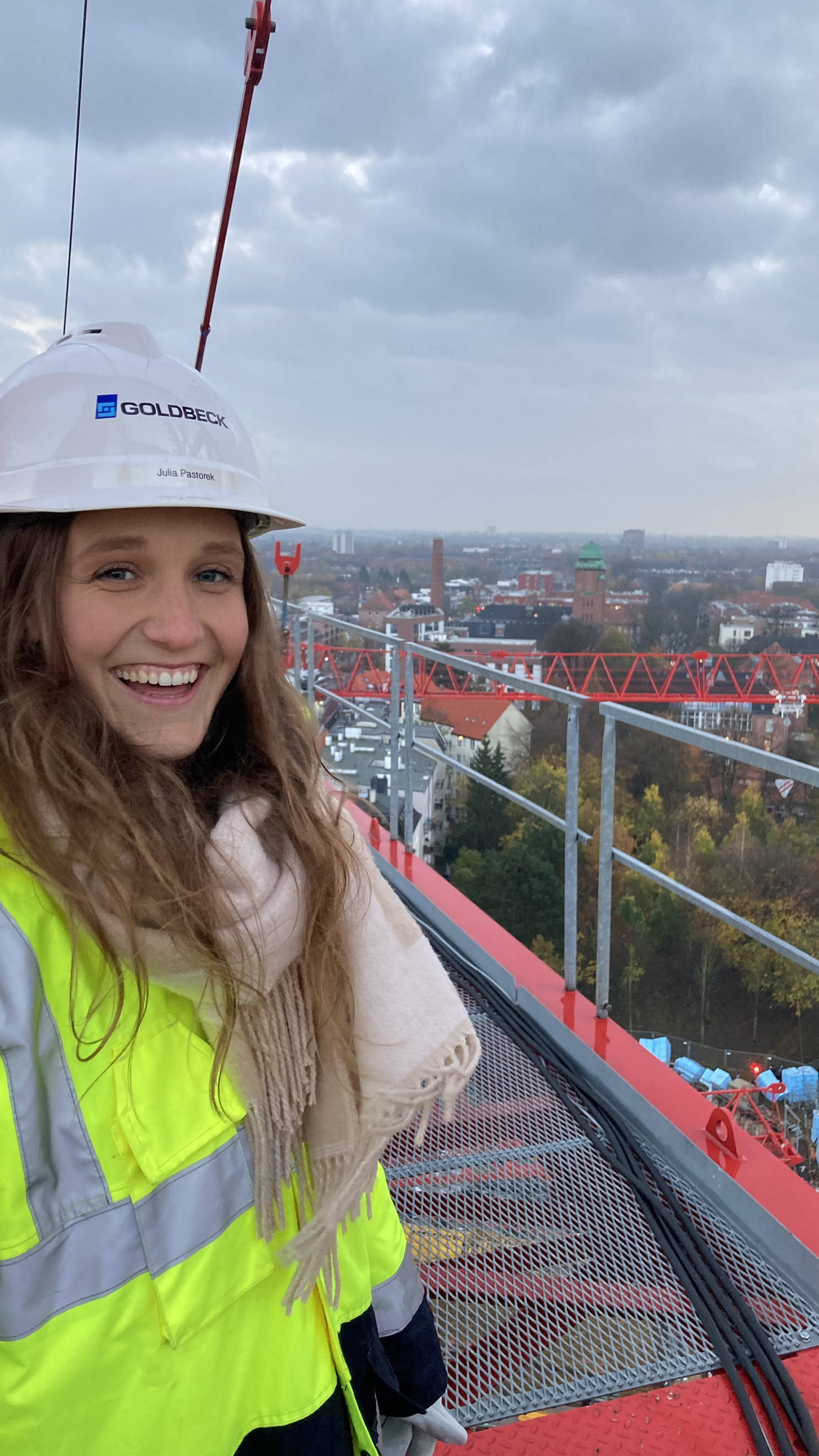 Frau in Sicherheitsweste und Helm steht lächelnd auf einer Baustelle mit Stadtlandschaft im Hintergrund.