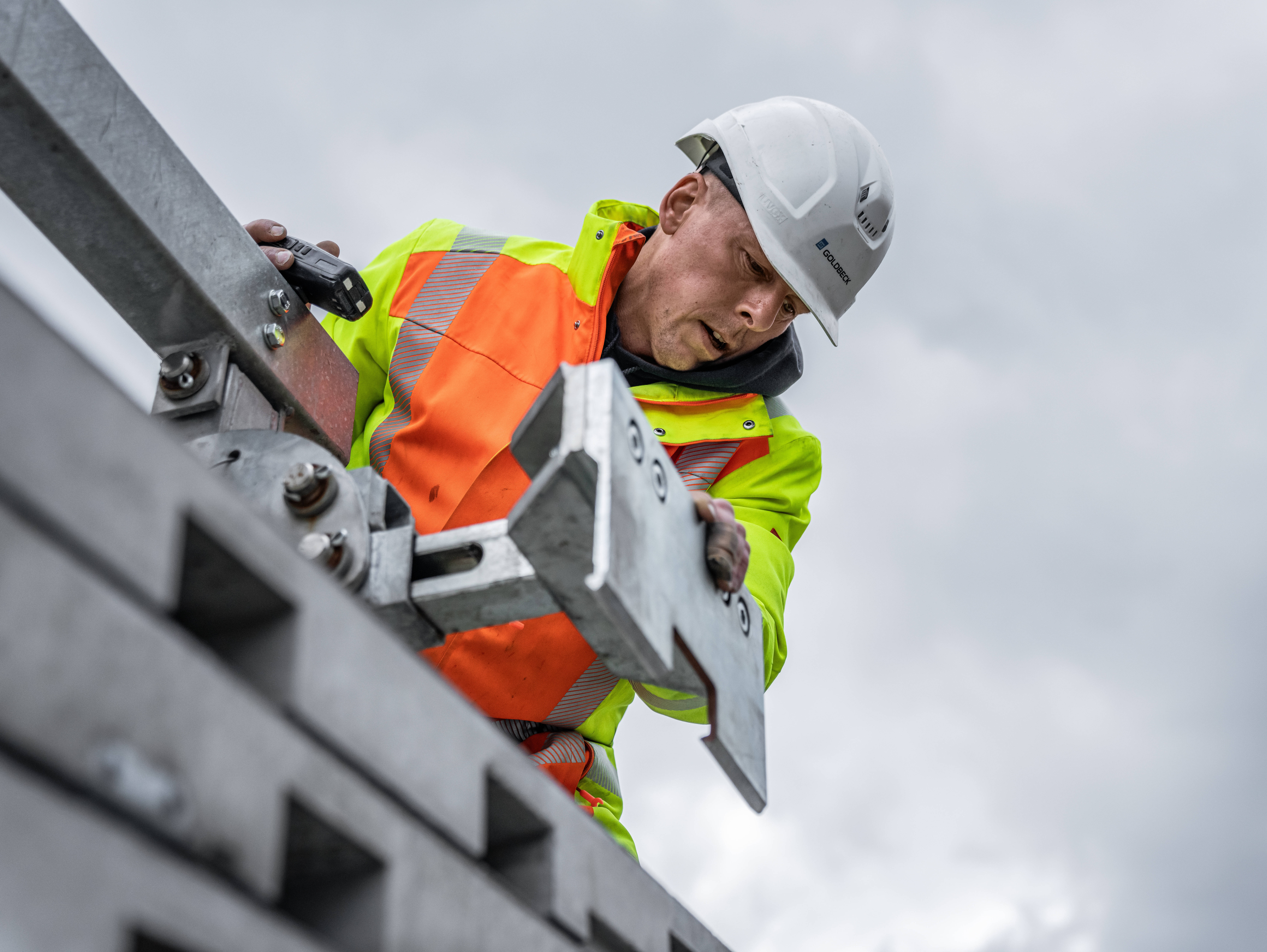 Bauarbeiter in Schutzkleidung bei der Arbeit an einer Metallkonstruktion unter bewölktem Himmel.