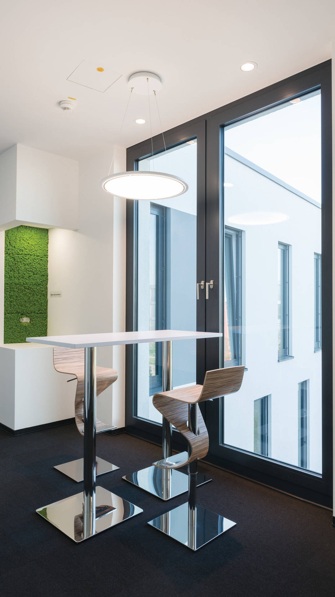 Moderner Besprechungsraum mit hohen Stühlen, einem weißen Tisch und großer Fensterfront, im Hintergrund eine begrünte Wand.