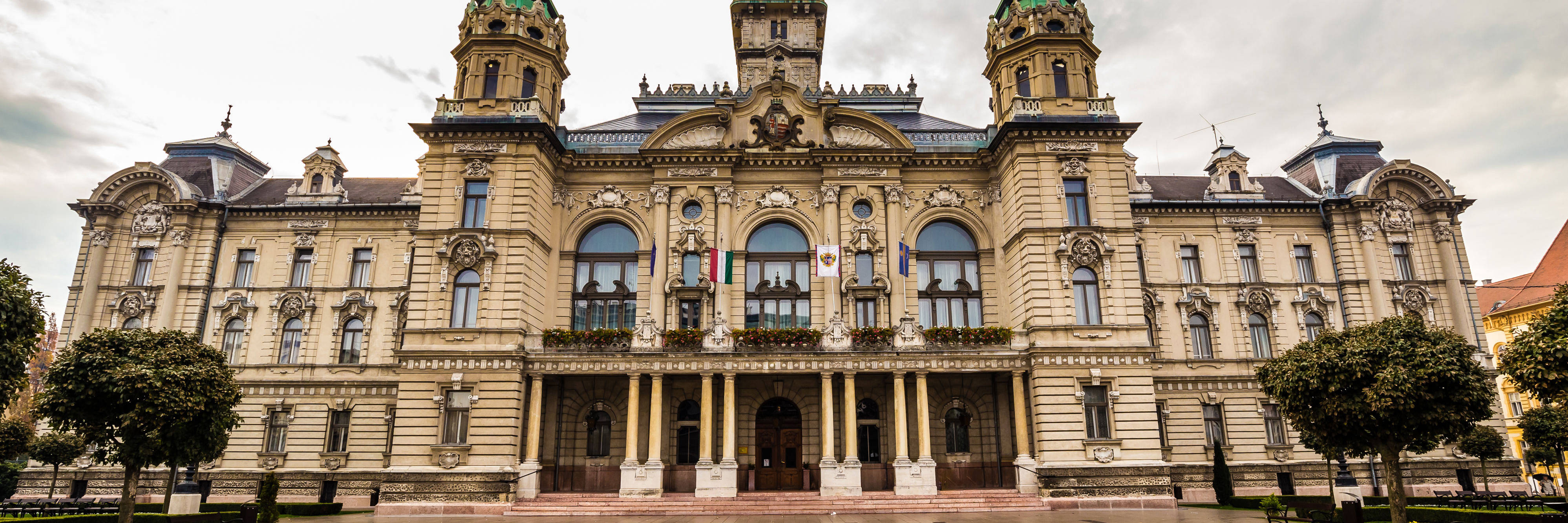 Historic building with towers and stylish details, front view of square with trees and overcast sky.