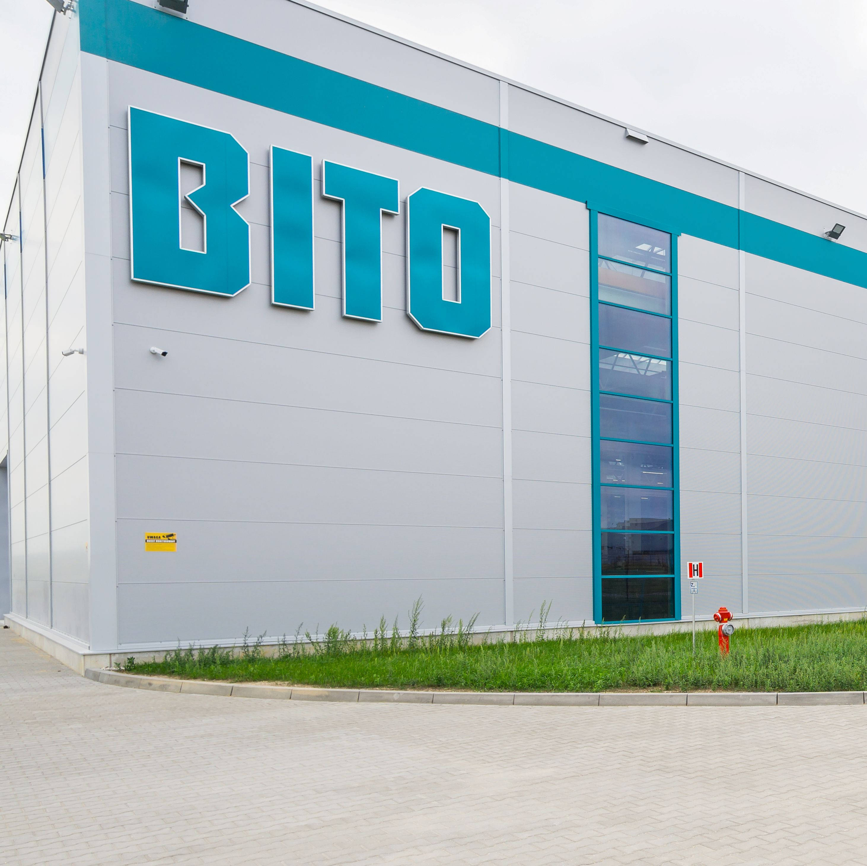 Nowoczesny budynek przemysłowy z logo "BITO" na fasadzie. Obiekt ma szare ściany z niebieskimi detalami i długie okna. Przed budynkiem znajduje się trawnik, a wokół biegnie brukowana droga. Na niebie widać pochmurne niebo.