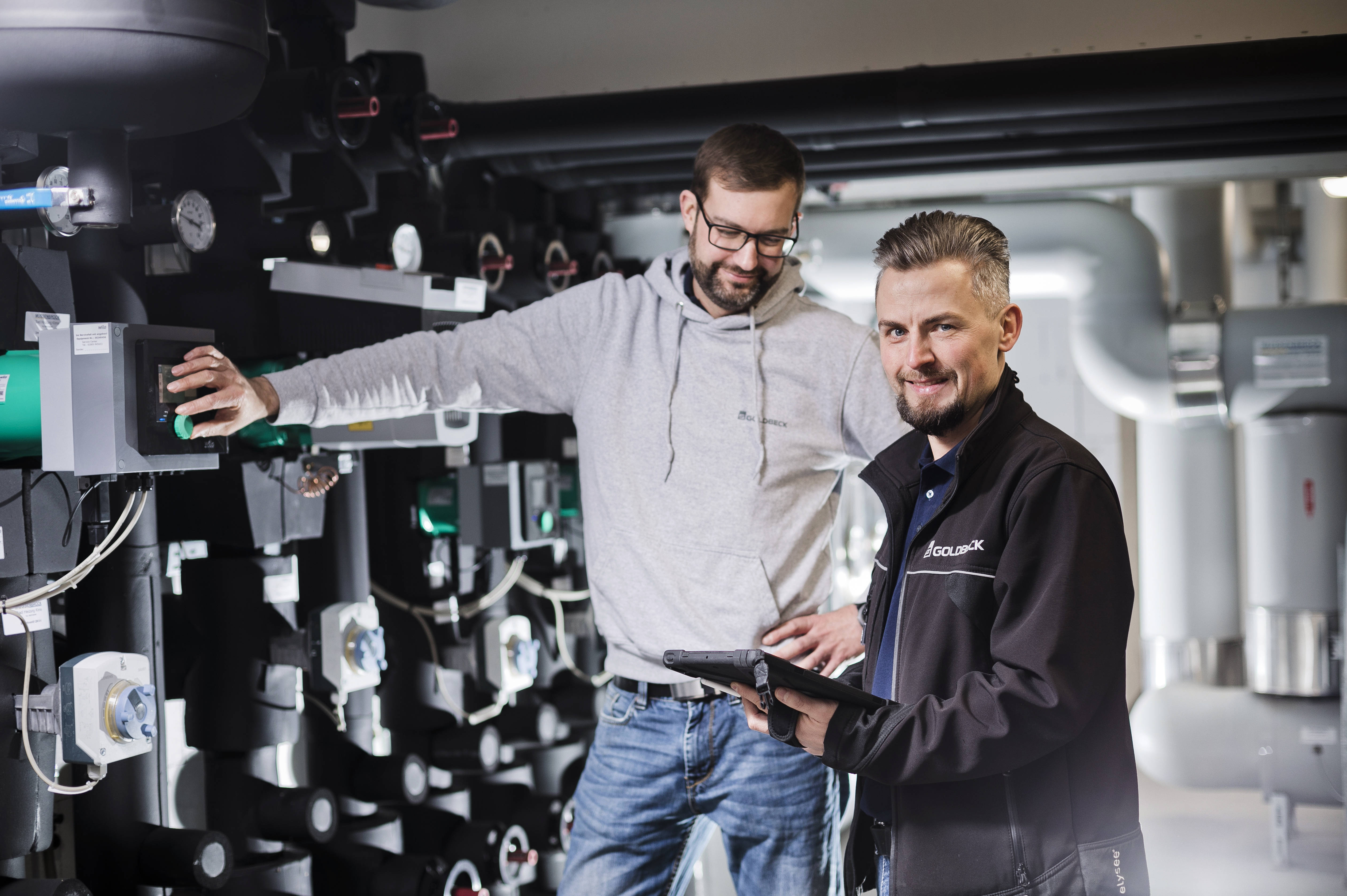 Two men in a technical facility, one operating a control panel, the other holding a tablet.