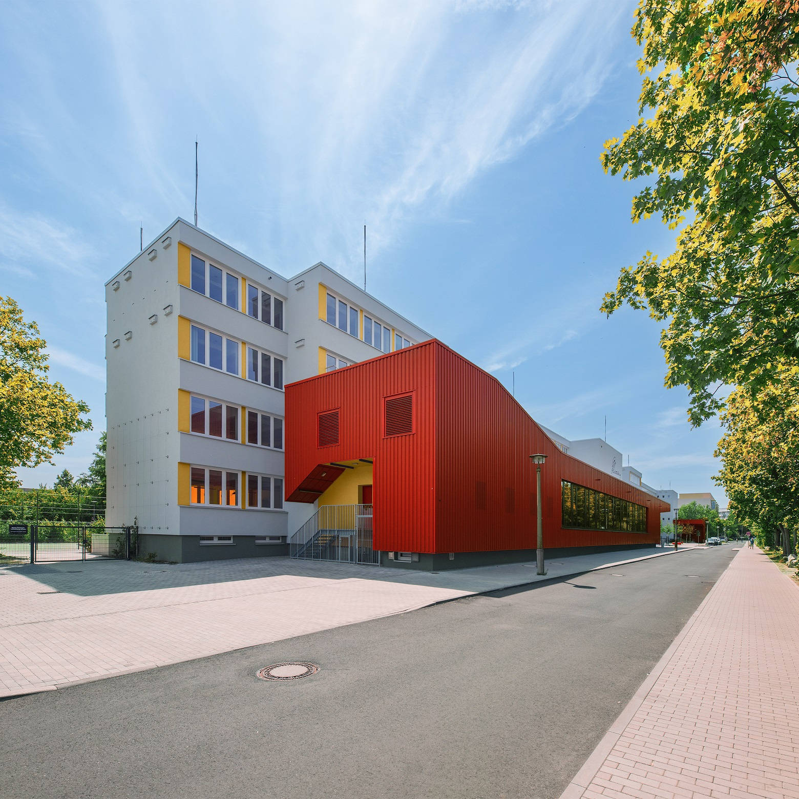 Moderner Schulbau mit weißer und roter Fassade, umgeben von Bäumen, unter klarem Himmel.