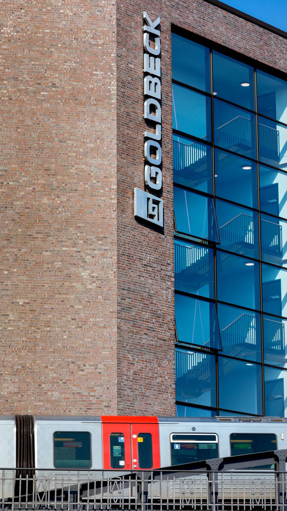 Moderne Glasfassade eines Bürogebäudes mit "GOLDBECK"-Logo, davor eine vorbeifahrende U-Bahn.