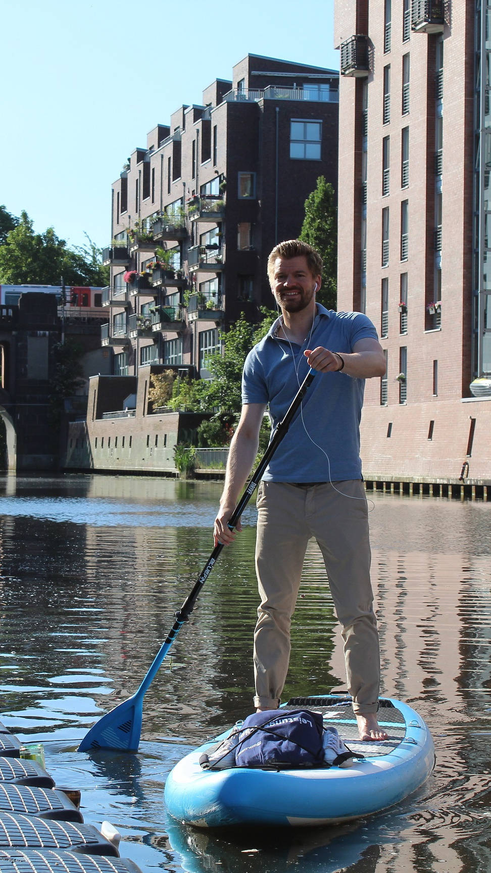 Mann steht auf Paddleboard, paddelt auf einem Kanal mit Brücke und modernen Backsteingebäuden im Hintergrund.