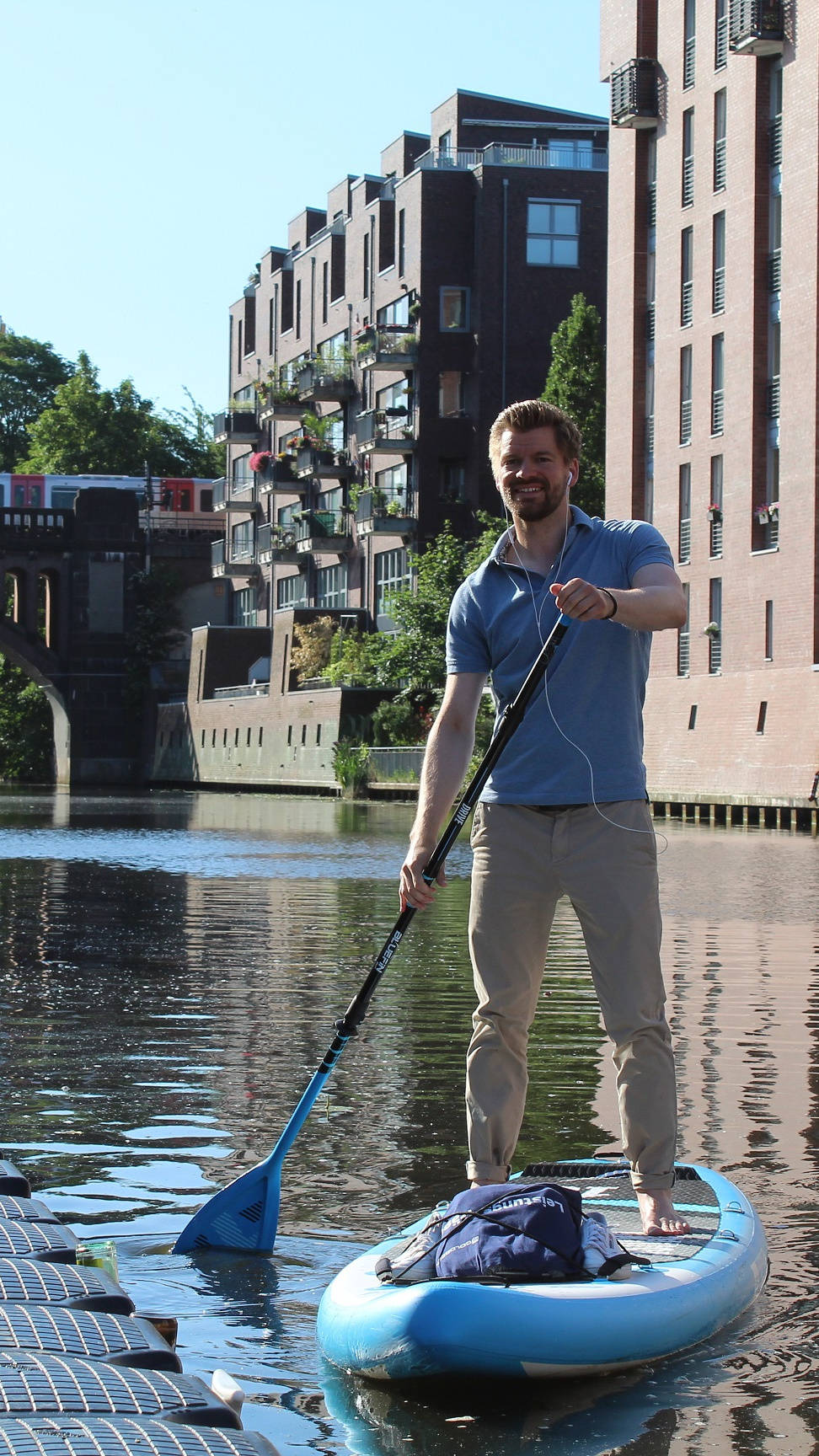 Mann steht auf Paddleboard, paddelt auf einem Kanal mit Brücke und modernen Backsteingebäuden im Hintergrund.