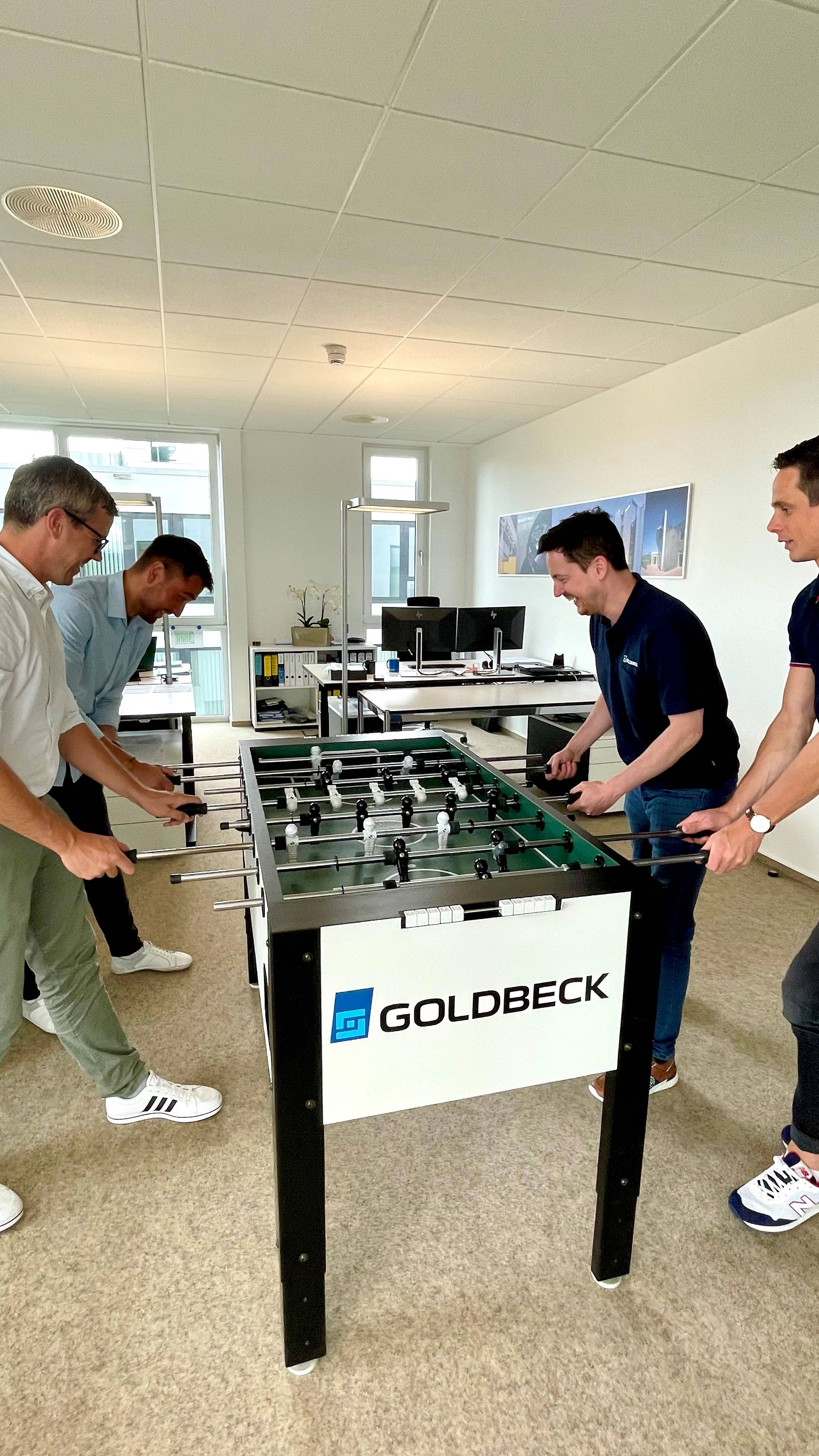 Mitarbeiter spielen Tischfußball in einem Büro, entspannen während einer Pause. Ein Tisch mit "Goldbeck"-Logo im Vordergrund.