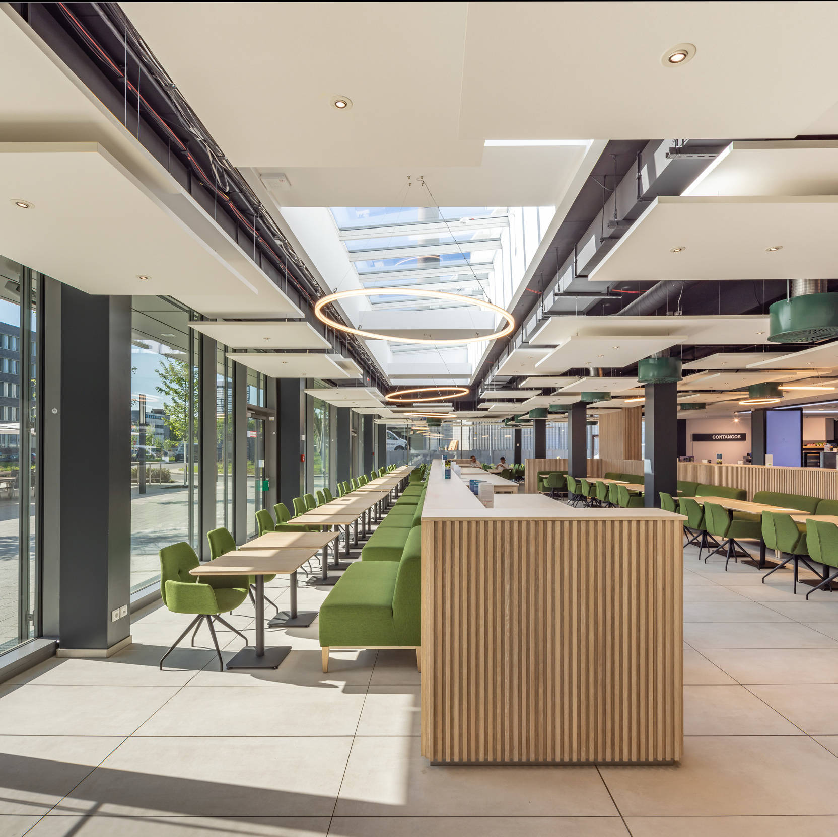 Modern, bright office café with green benches and tables, large window and circular hanging lamps.