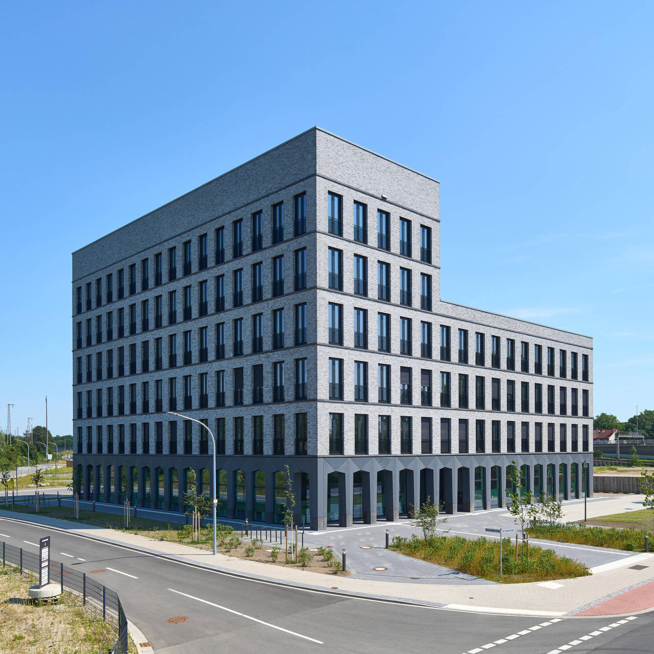 Modernes, sechsstöckiges Bürogebäude aus grauem Backstein an einer Straße, umgeben von Grünflächen, unter klarem blauem Himmel.