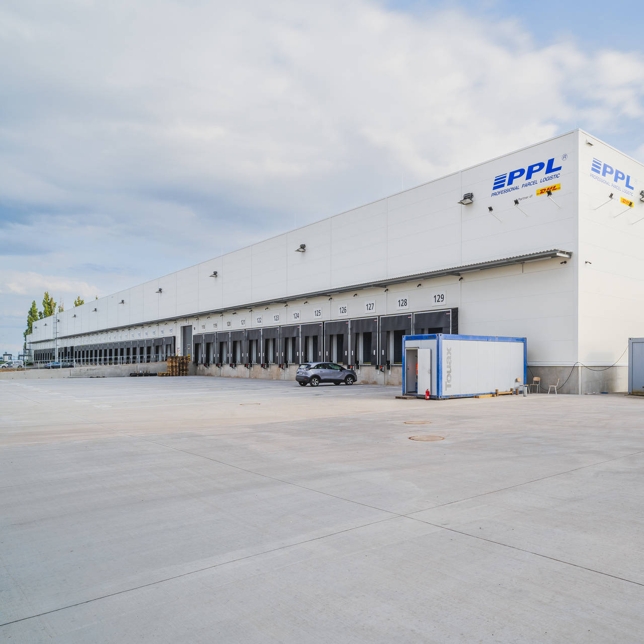 Distribuční centrum s několika nakládacími rampami a markýzou, vozidla zaparkovaná na prostorném parkovišti.
