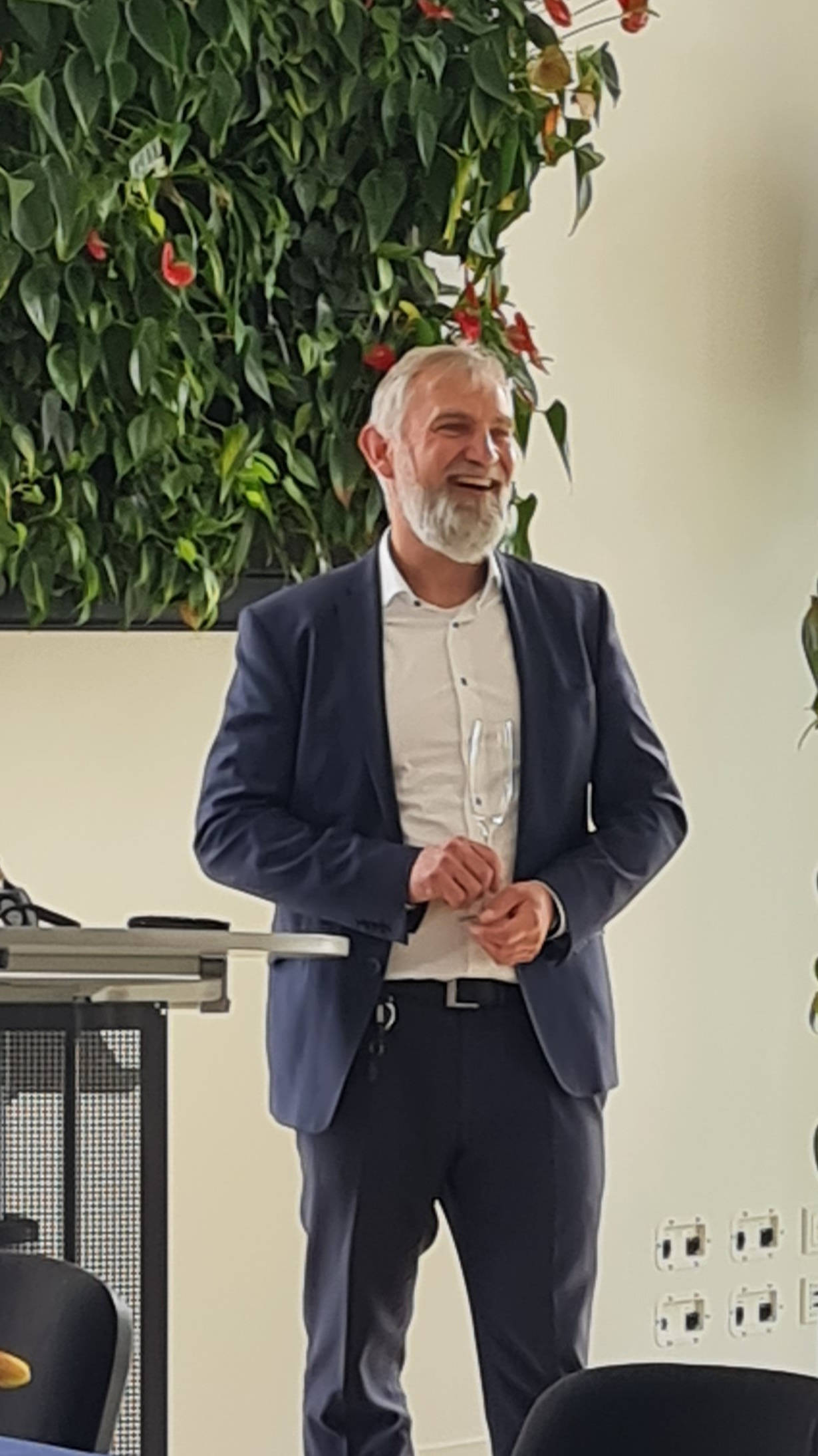 Mann im Anzug lächelt bei einer Veranstaltung neben einem Podium, mit grüner Pflanzenwand im Hintergrund.