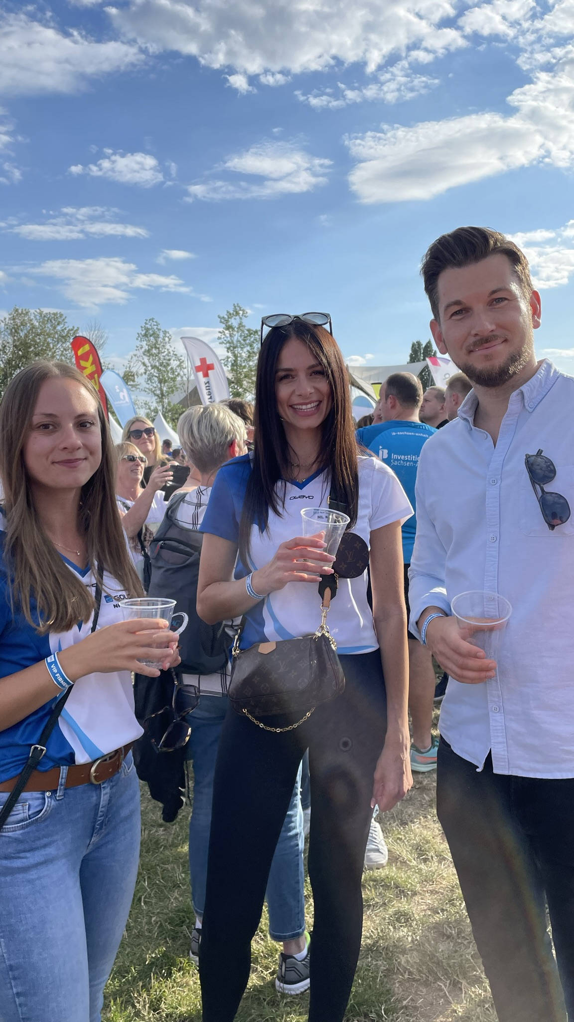 Gruppe von drei Personen bei einer Outdoor-Veranstaltung, lächelnd und Getränke haltend, mit blauem Himmel und Menschenmenge im Hintergrund.