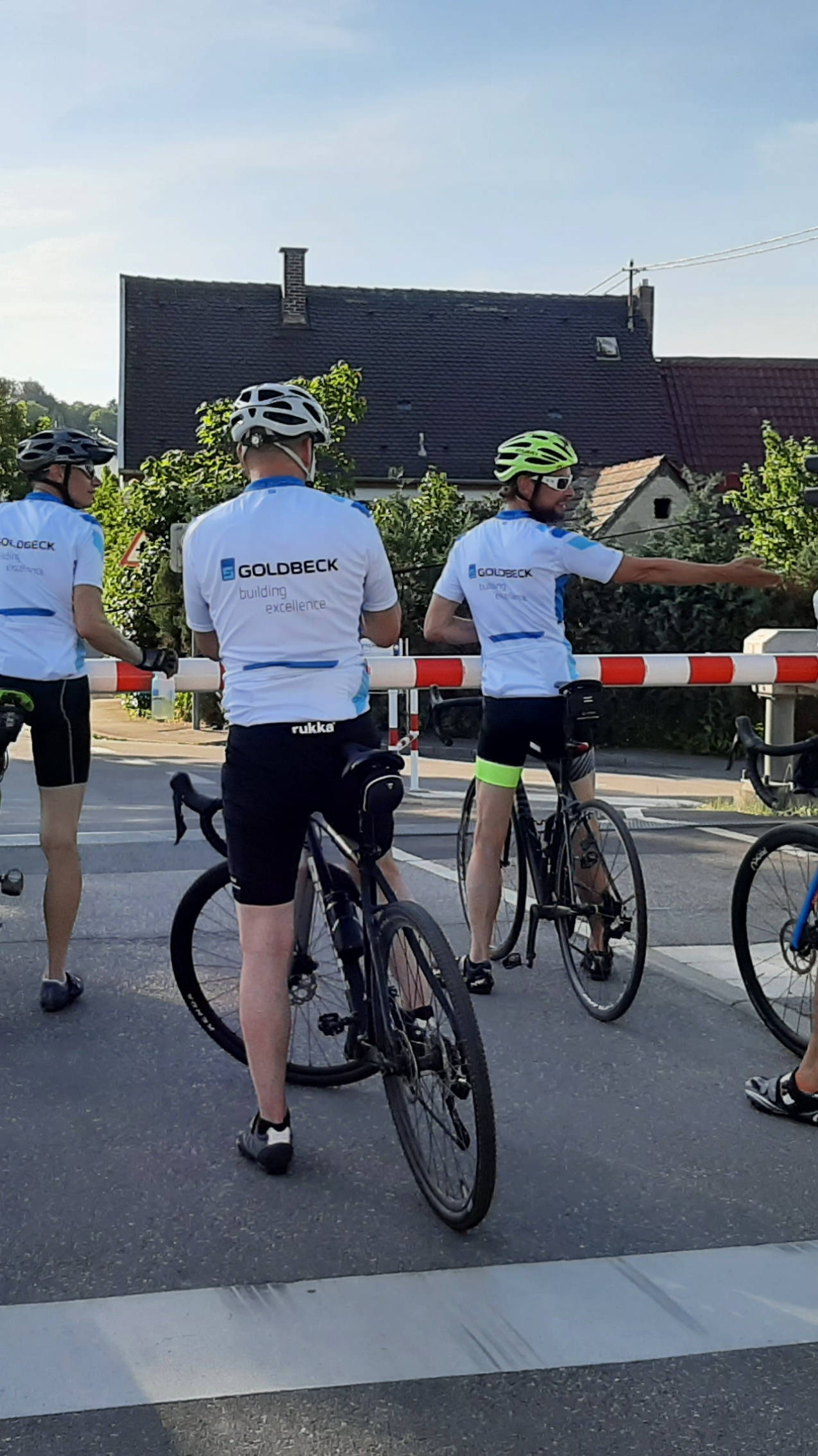 Fünf Radfahrer in Sportkleidung warten an einem geschlossenen Bahnübergang bei roter Ampel.