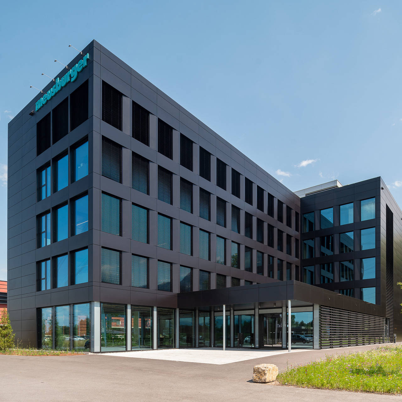 Modernes Bürogebäude mit Glasfassade unter blauem Himmel, umgeben von Grünflächen.