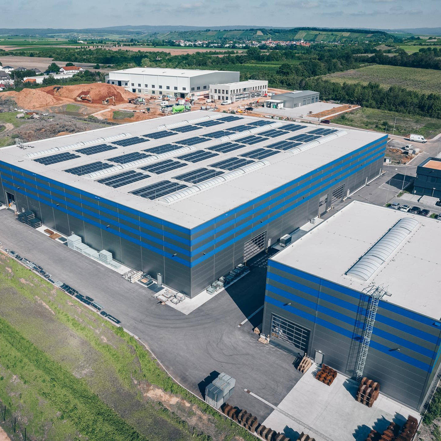 Luftaufnahme eines modernen Industriegebäudes mit Solaranlagen auf dem Dach, umgeben von grüner Landschaft und weiteren Gebäuden im Hintergrund.