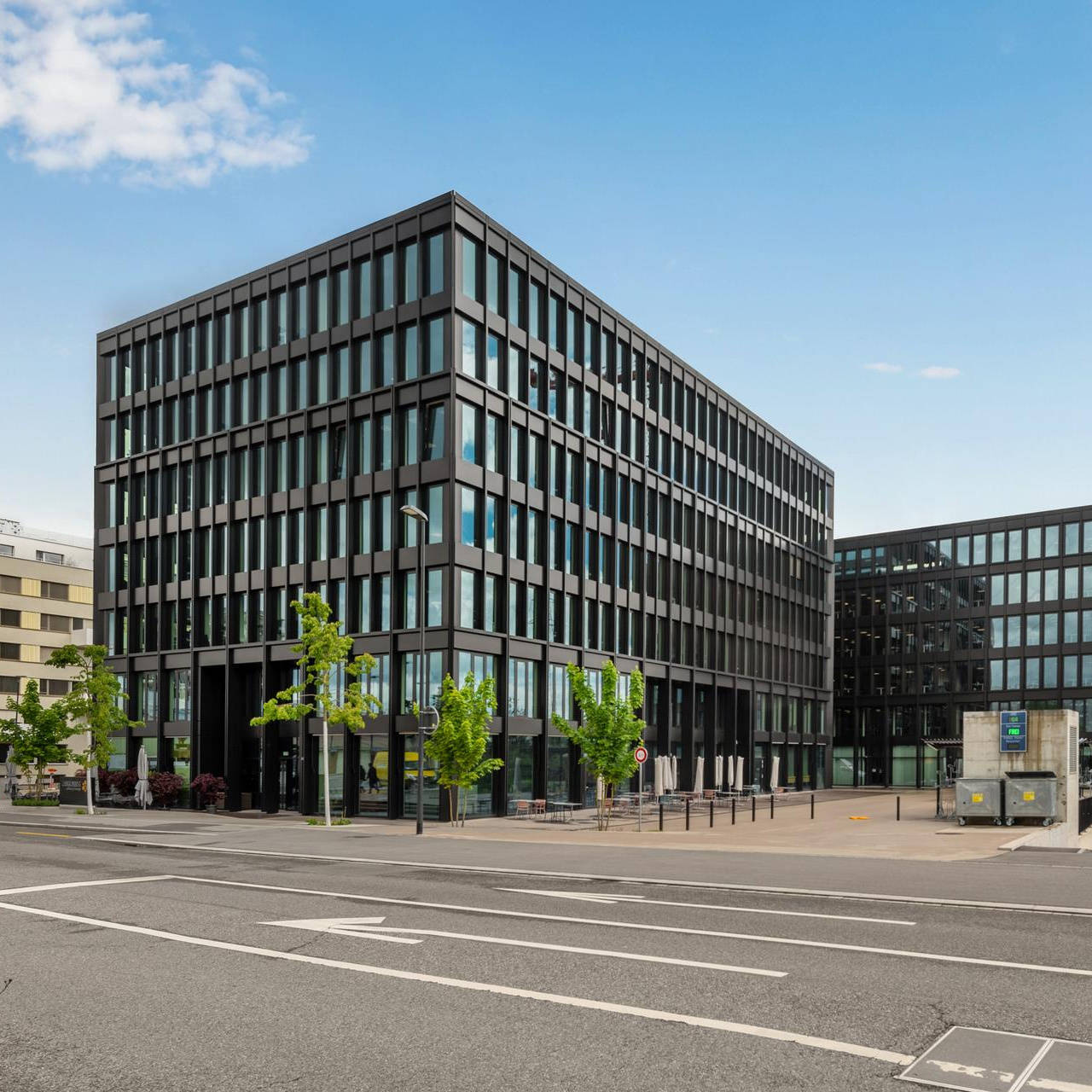 Moderne Bürogebäude mit Glasfassade bei blauem Himmel, umgeben von Straßenkreuzung und grünen Bäumen.