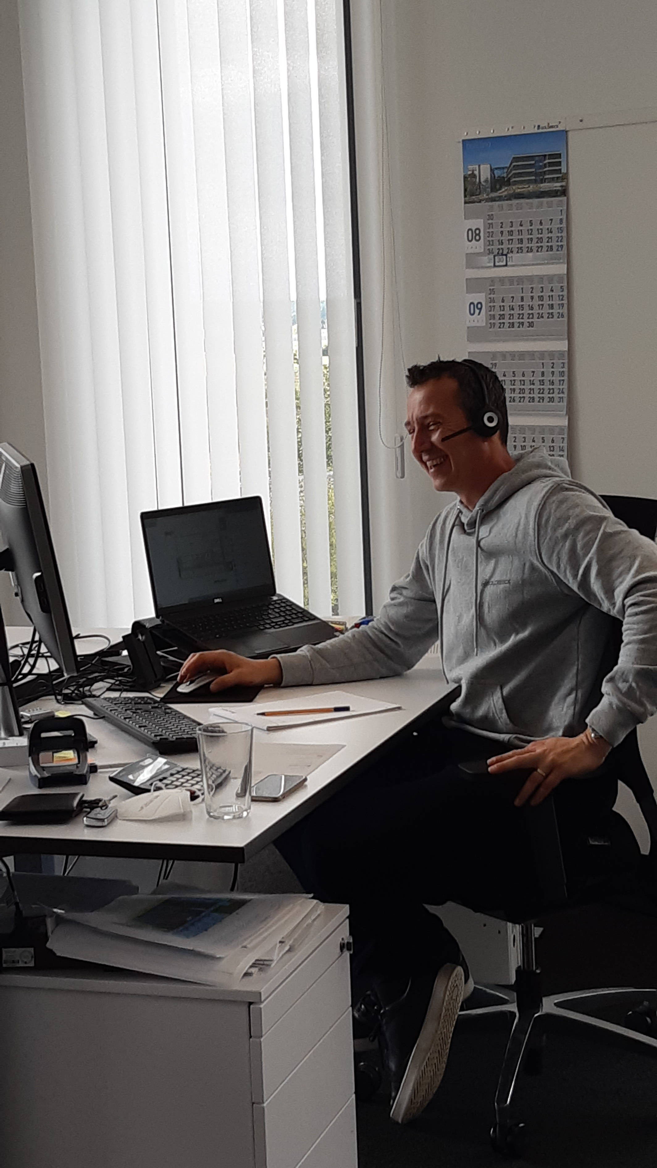 Person am modernen Schreibtisch, mit einem Headset telefonierend, lächelnd, vor Computerbildschirm sitzend. Büro mit Kalender im Hintergrund.