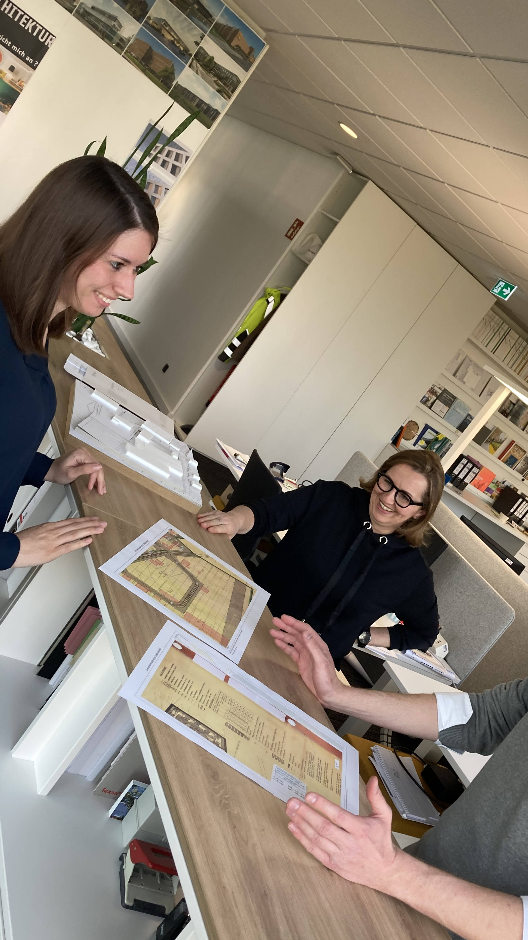 Drei Personen diskutieren an einem Schreibtisch in einem Büro über Baupläne.