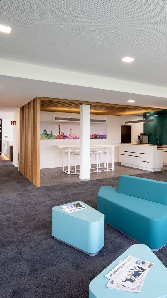 Modern lounge with blue seating, coffee table with newspapers and kitchen in the background, decorated with a skyline painting.