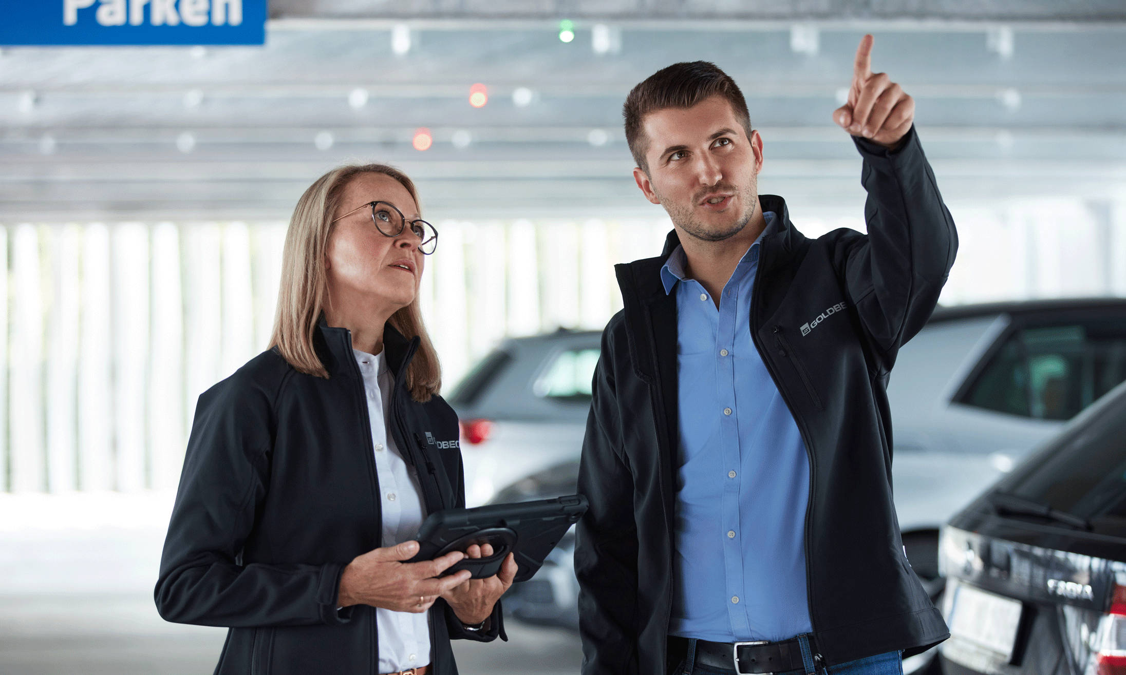 Man and woman in underground parking garage, woman holding a tablet, man pointing to a parking guidance system sign.