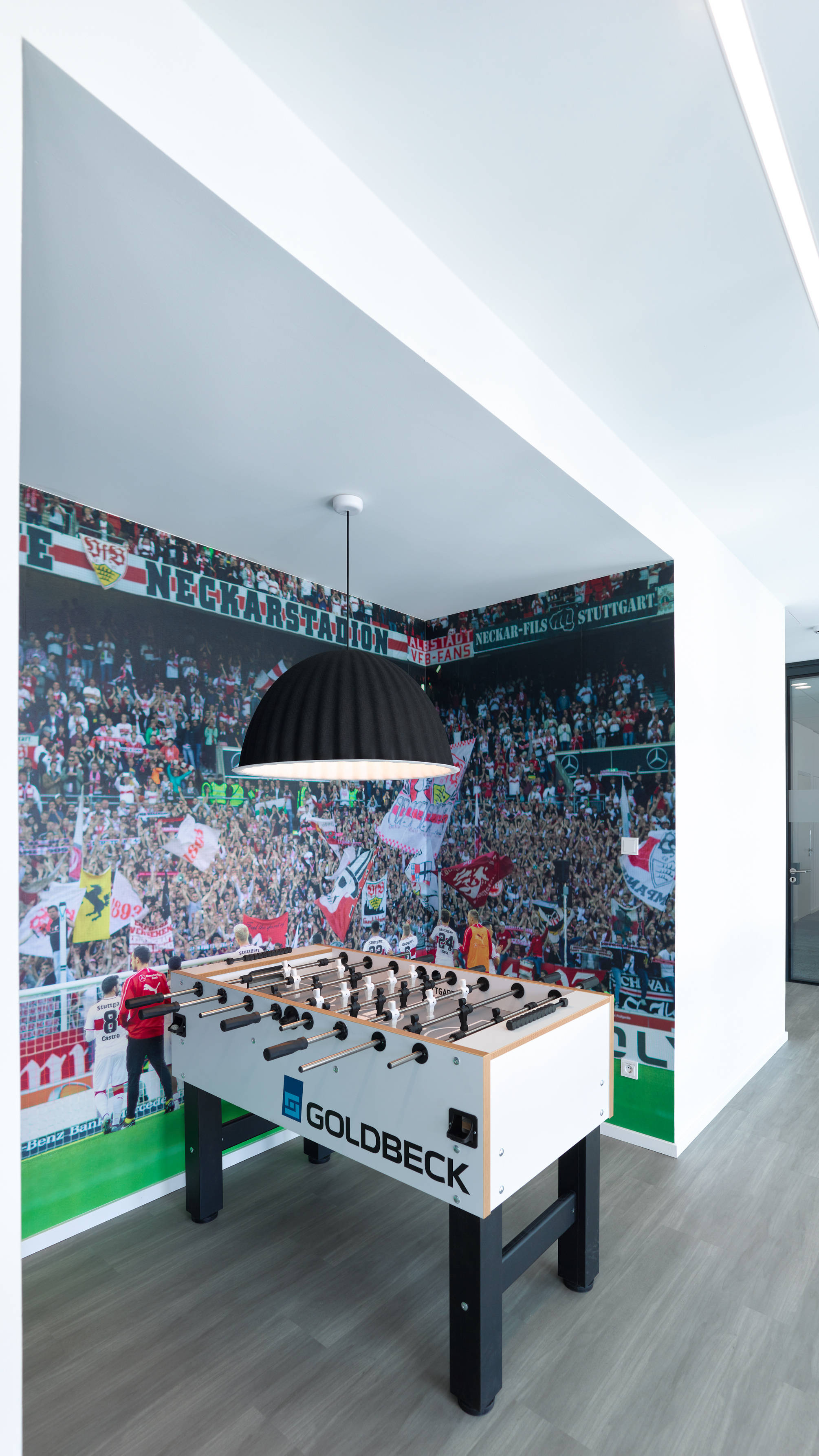 Tischkicker in modernem Raum, umgeben von Fußballstadion-Wandbildern, mit Deckenleuchte darüber.