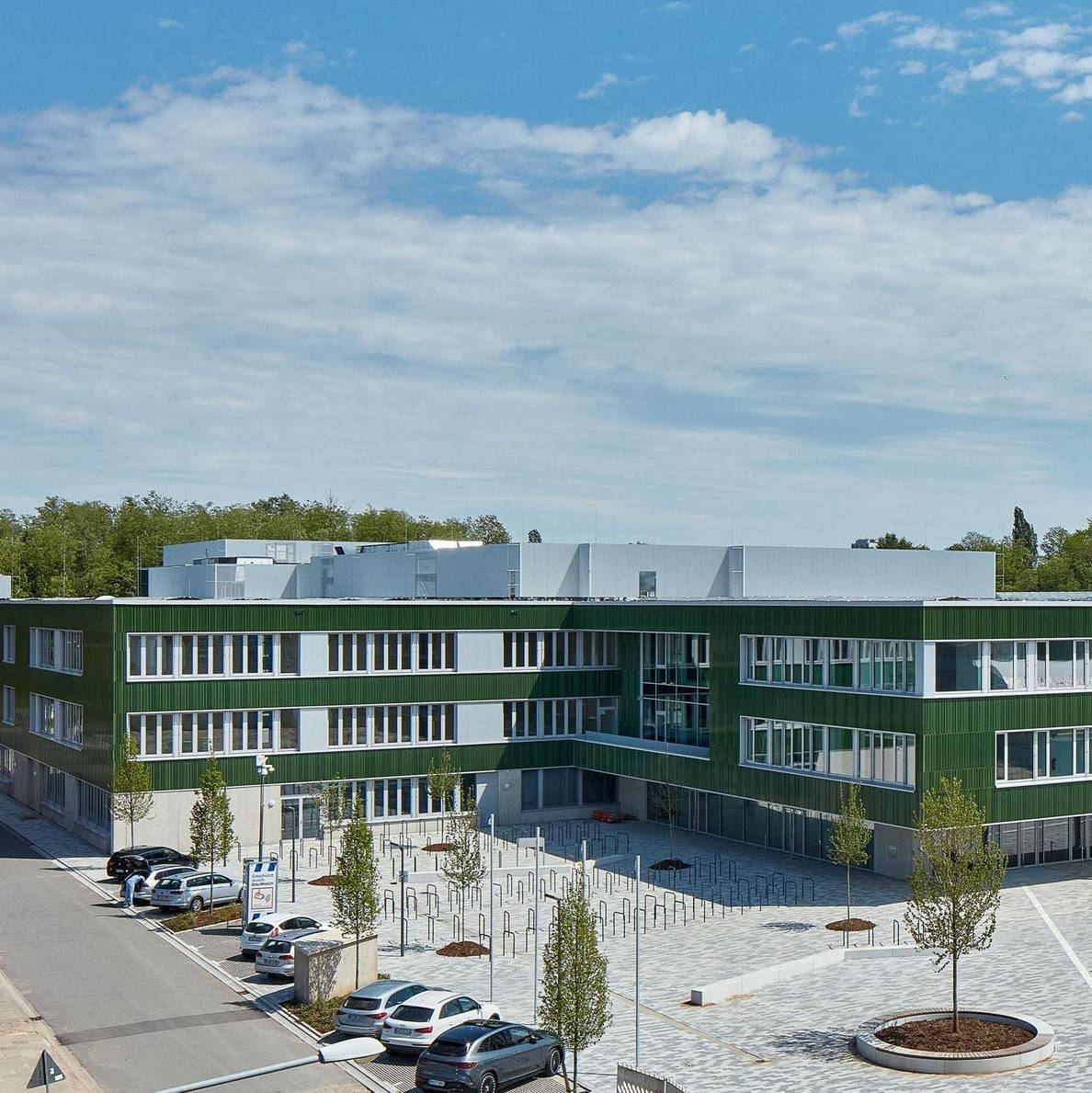 Moderne, grün-weiße Schulgebäude mit großem gepflastertem Vorplatz, Bäumen und geparkten Autos an einem sonnigen Tag.