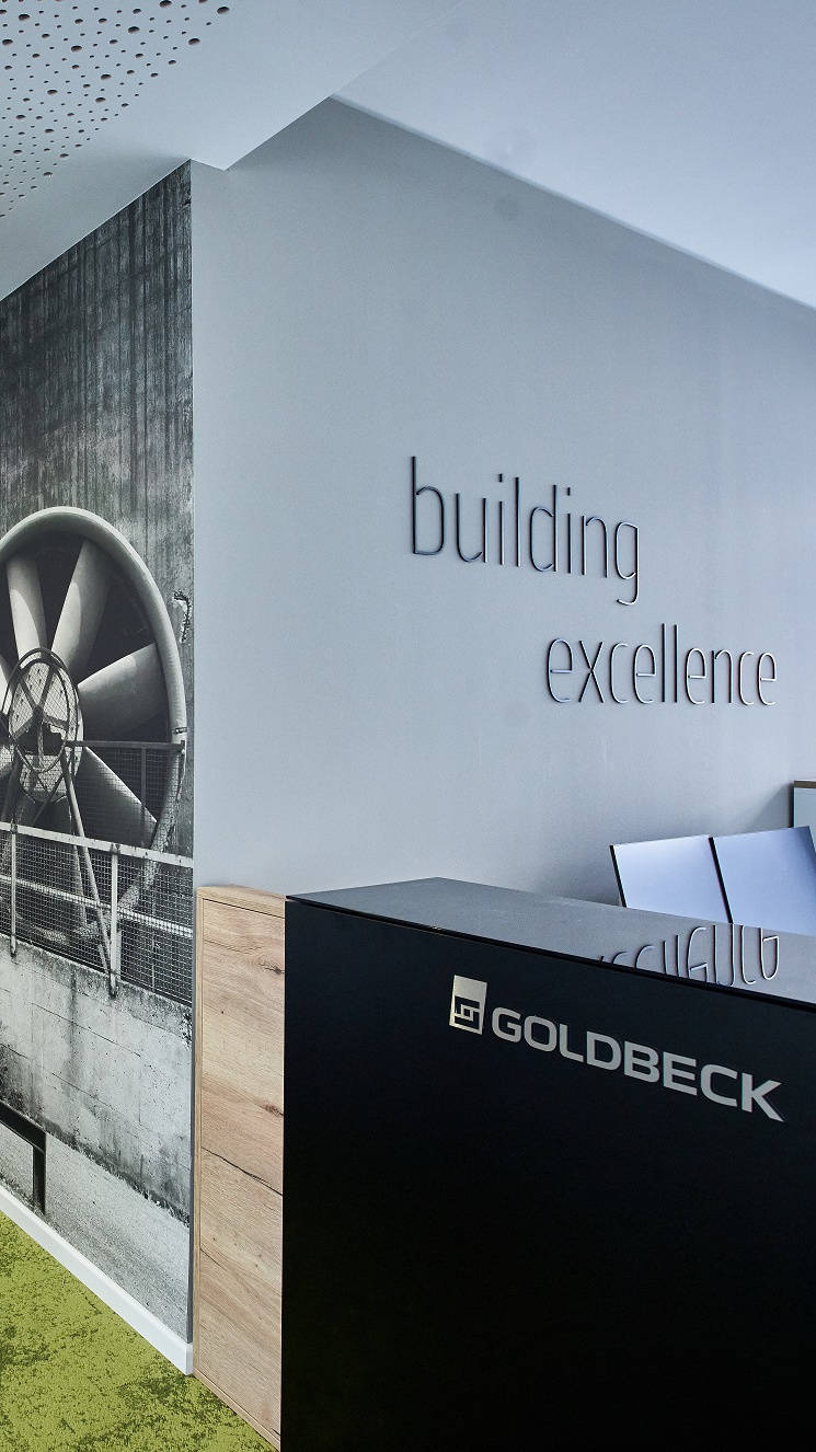 Moderner Büroflur mit geschwungenem Design und Grünpflanzen, Wandkunst mit Ventilator-Motiv und "building excellence"-Schriftzug.