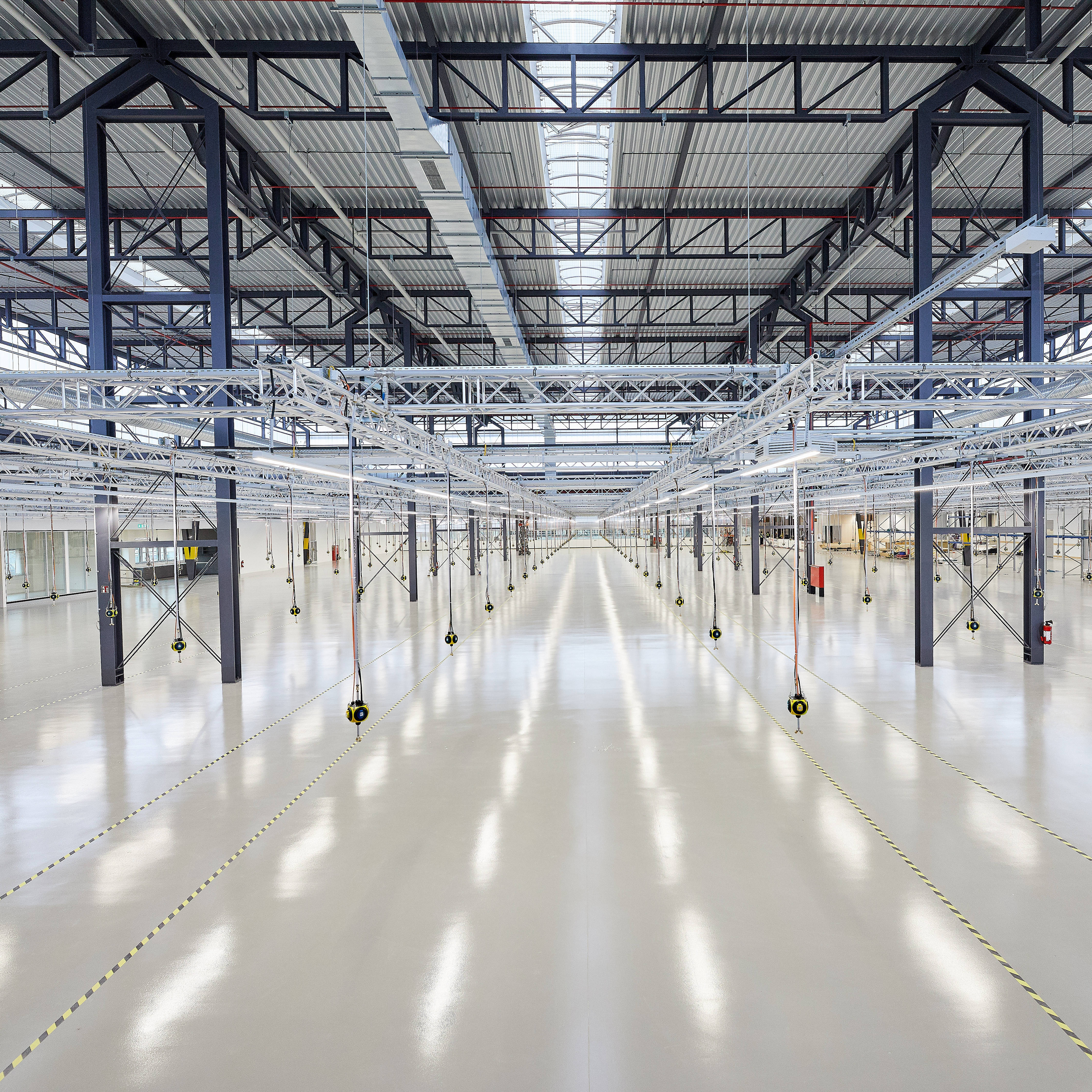Große, leere Industriehalle mit hoher Decke und Oberlichtern, umgeben von Stahlträgern und Beleuchtungseinrichtungen.