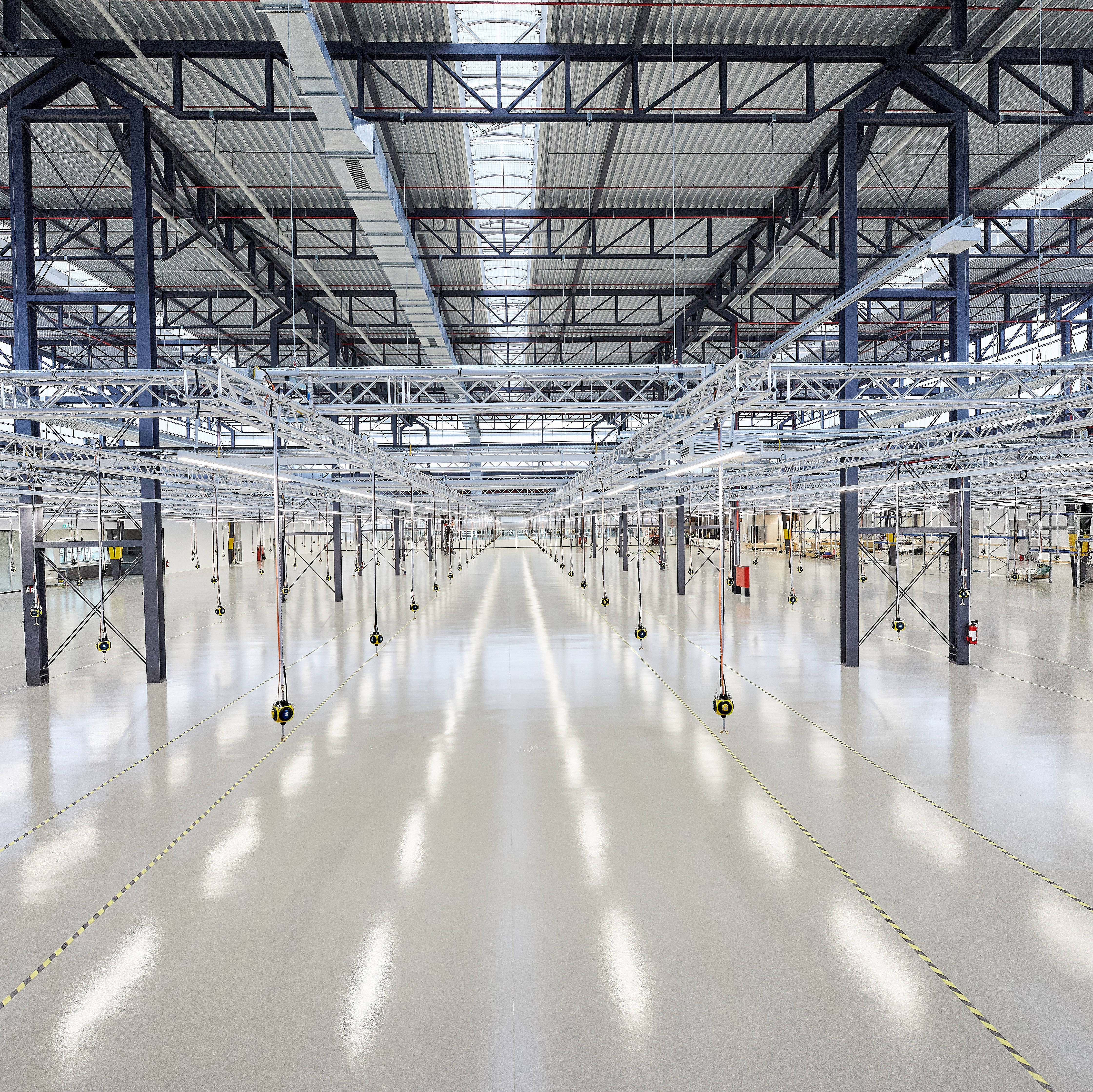Große, leere Industriehalle mit hoher Decke und Oberlichtern, umgeben von Stahlträgern und Beleuchtungseinrichtungen.