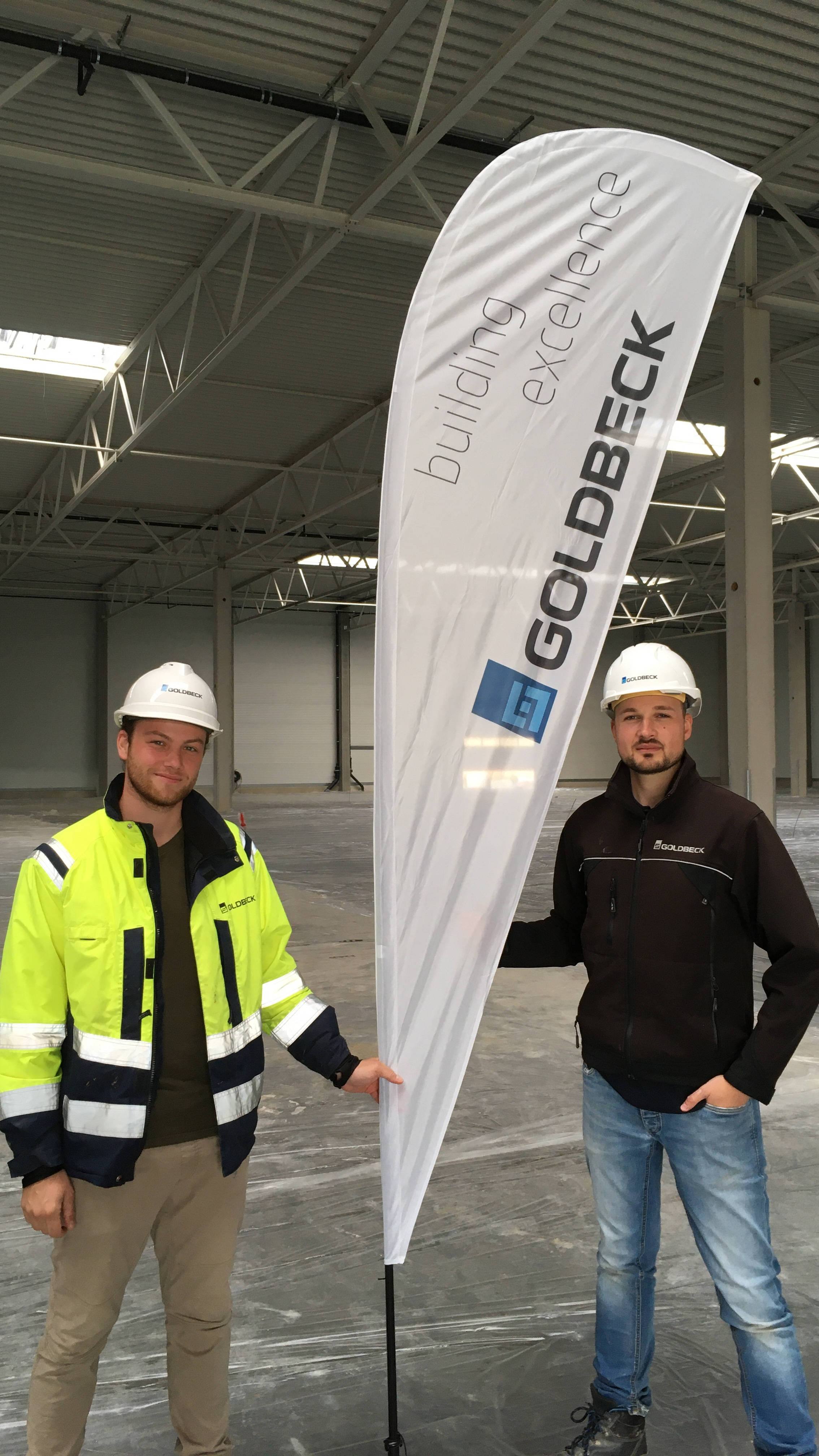 Zwei Bauarbeiter in Schutzkleidung halten ein Werbebanner in einer großen Halle, das "Goldbeck" zeigt.