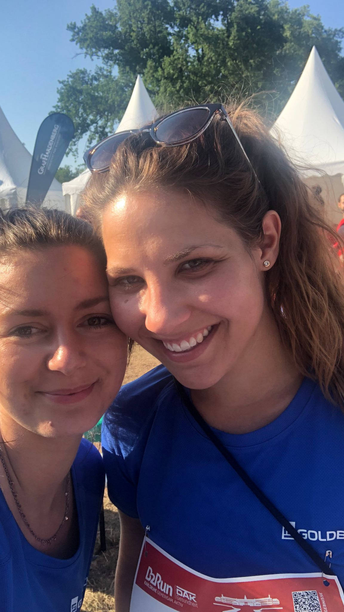 Zwei Frauen in blauen Laufshirts lächeln bei einem Outdoor-Sportevent, im Hintergrund sind weiße Zelte zu sehen.