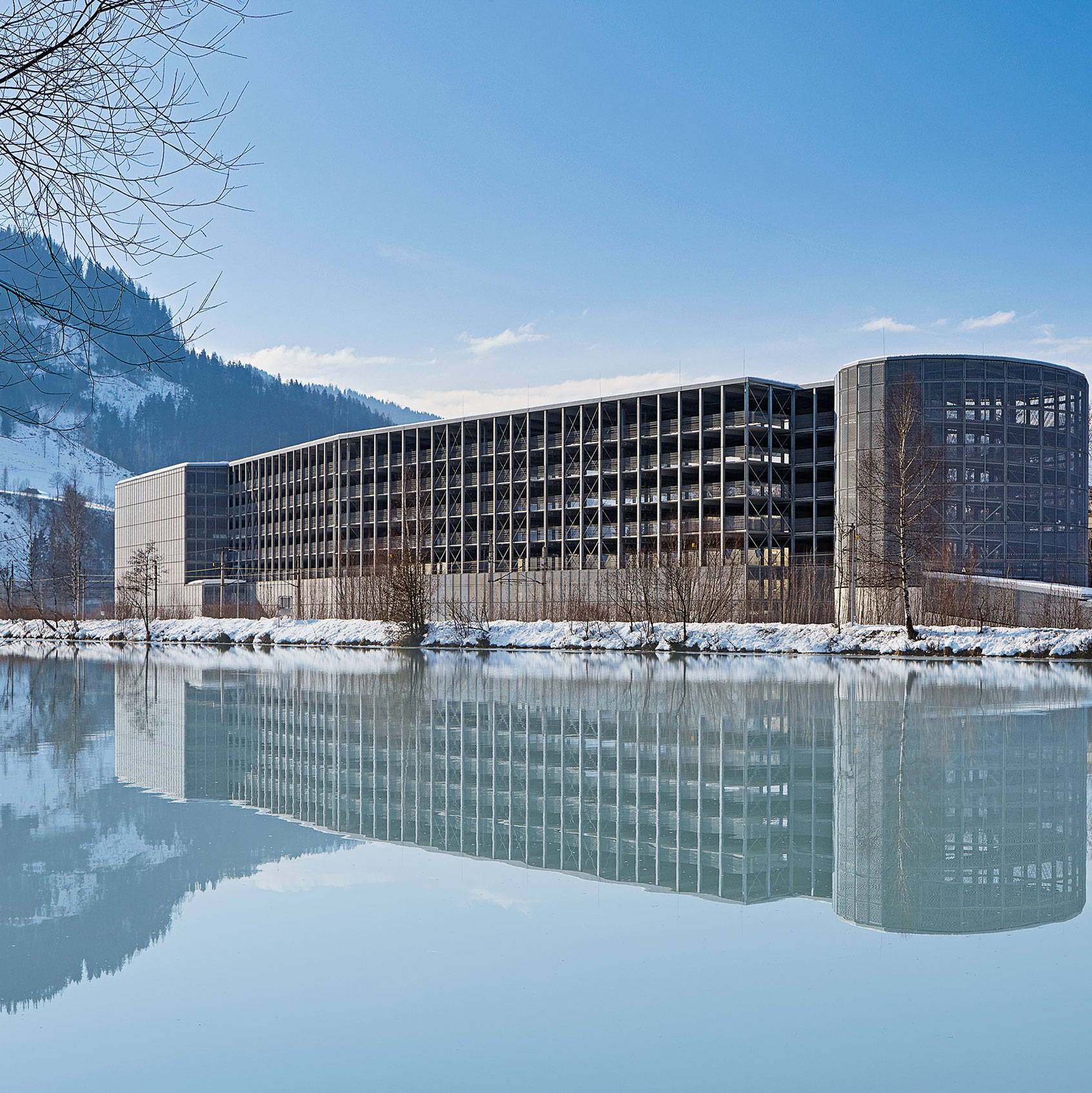 Moderner Gebäudekomplex am See, umgeben von schneebedeckten Bergen und Bäumen, spiegelt sich im Wasser wider.