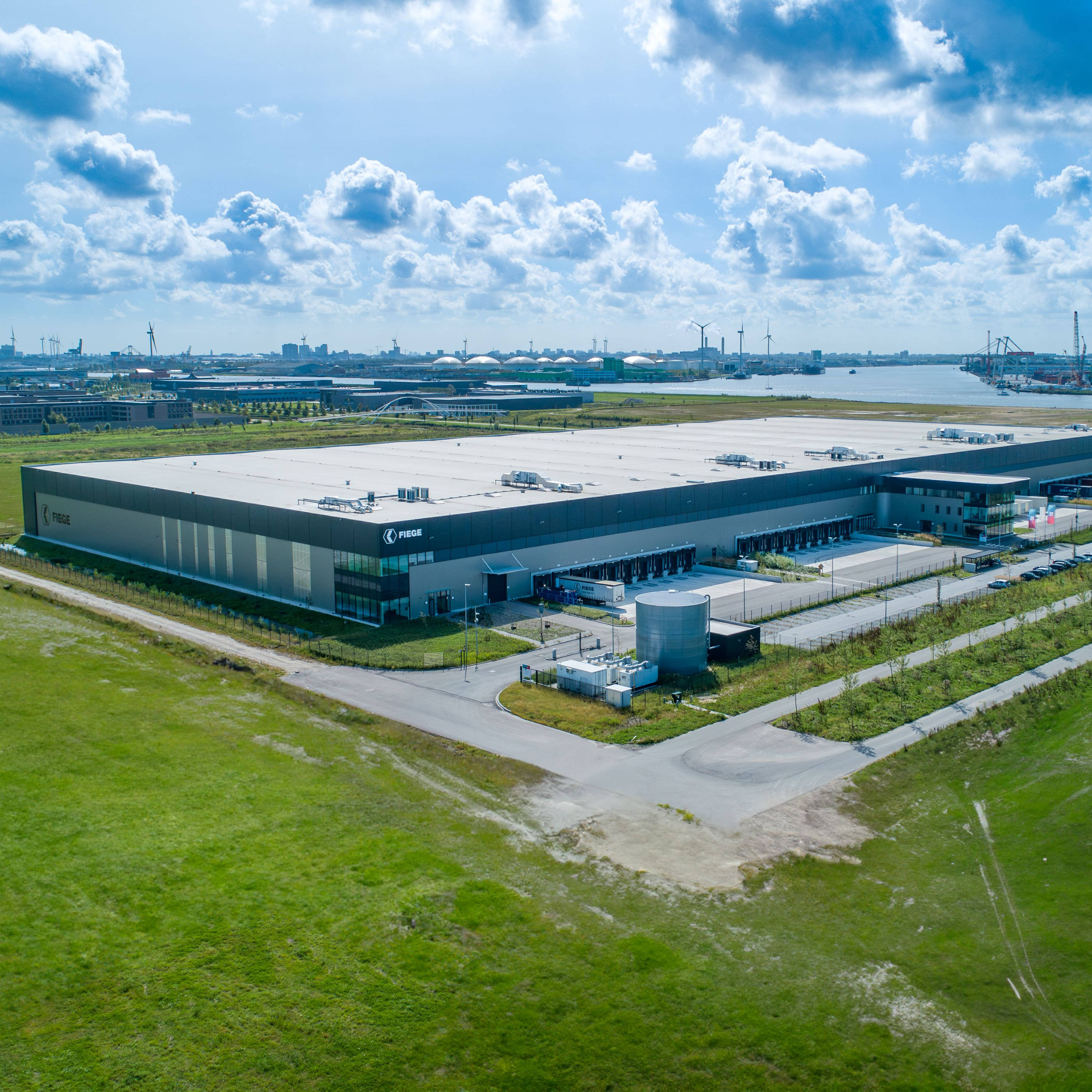 Luchtfoto van een groot distributiecentrum omringd door groen grasland, met op de achtergrond een waterweg en industriële structuren, waaronder kranen en windturbines. De lucht is deels bewolkt met verspreide wolken.