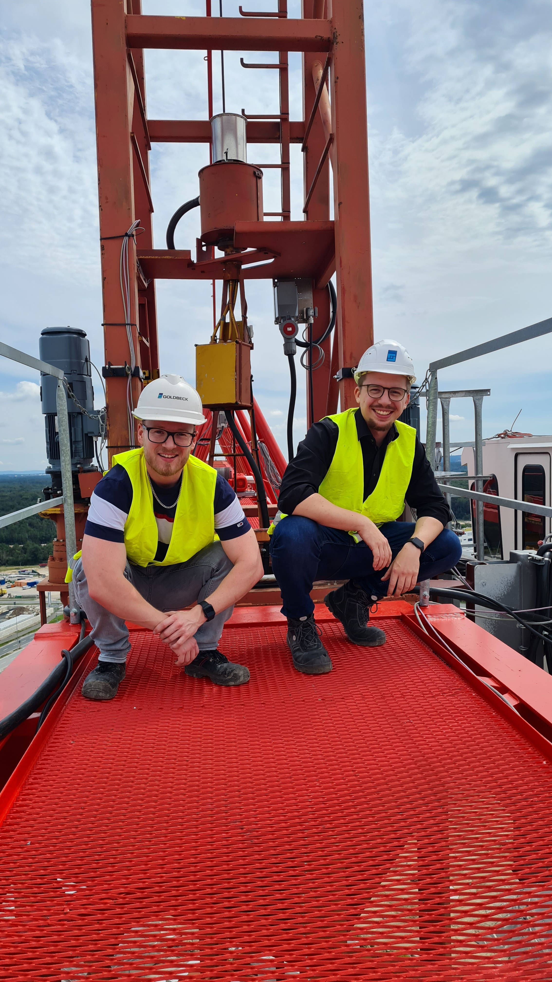 Zwei Bauarbeiter in gelben Warnwesten und Helmen knien auf einem roten Baugerüst mit Aussicht über eine Baustelle.