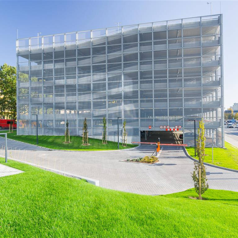 Nowoczesny, wielopoziomowy parking wyłożony szarą siatką, z wjazdem od frontu. Przed budynkiem znajduje się zielony trawnik oraz młode drzewa. W tle widać ulice i budynki miejskie.