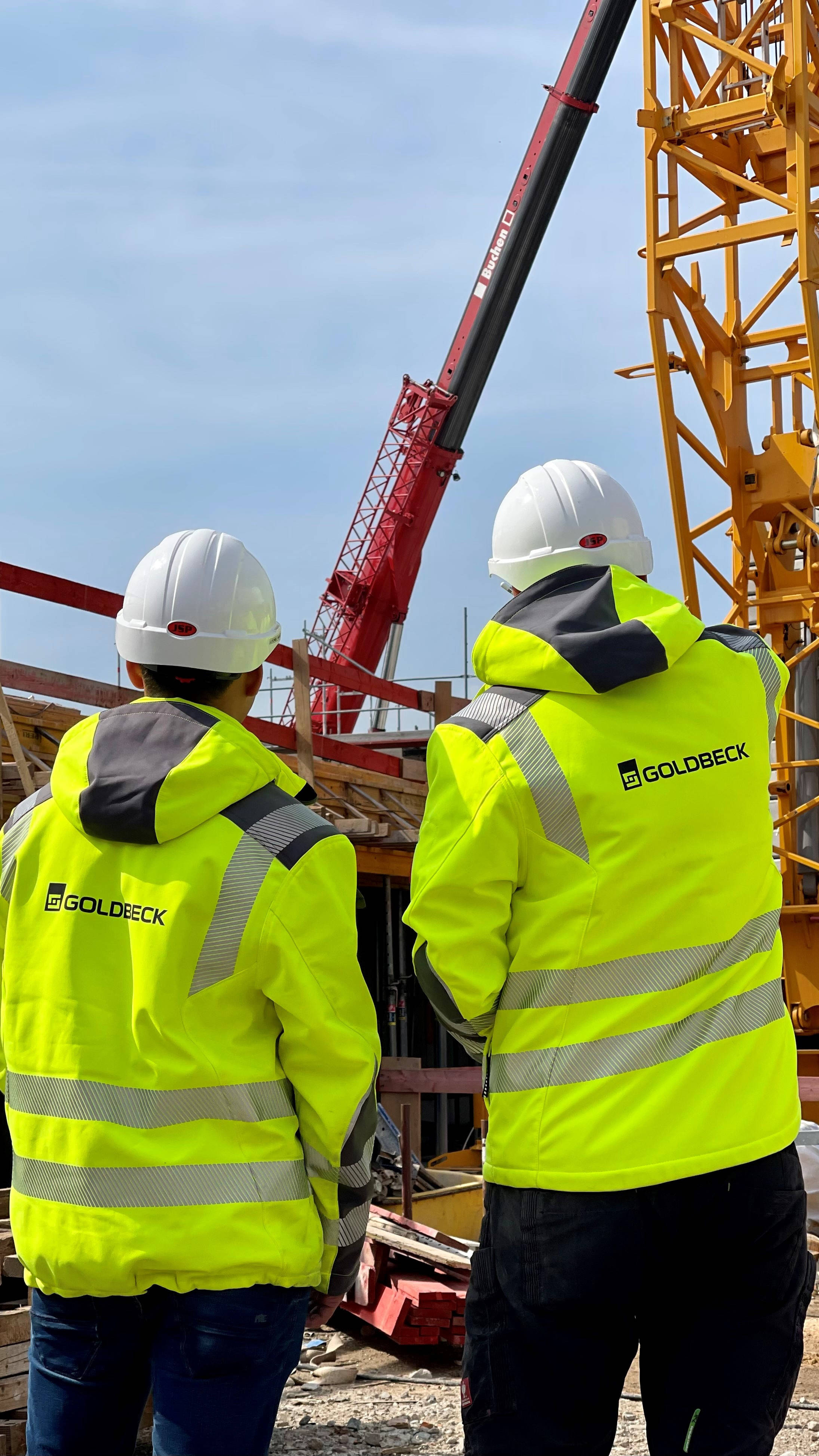 Zwei Bauarbeiter in gelben Sicherheitsjacken und Helmen betrachten eine Baustelle mit Kran im Hintergrund.
