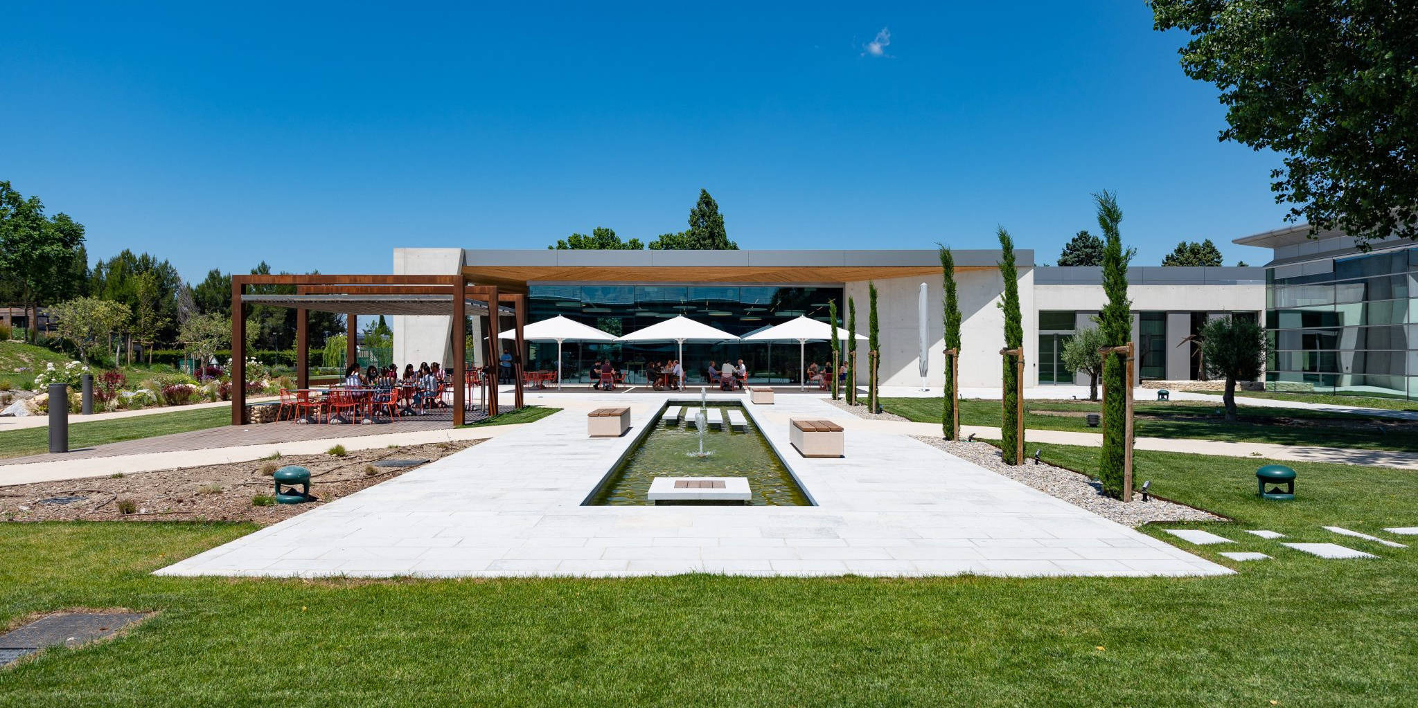 Ein moderner Gebäudekomplex mit einer klaren Glasfront und einer einladenden Terrasse, gestaltet von Goldbeck. Der grüne Außenbereich mit makellos gepflegtem Rasen und einem dekorativen Wasserbecken schafft eine harmonische Verbindung zwischen Architektur und Landschaft.