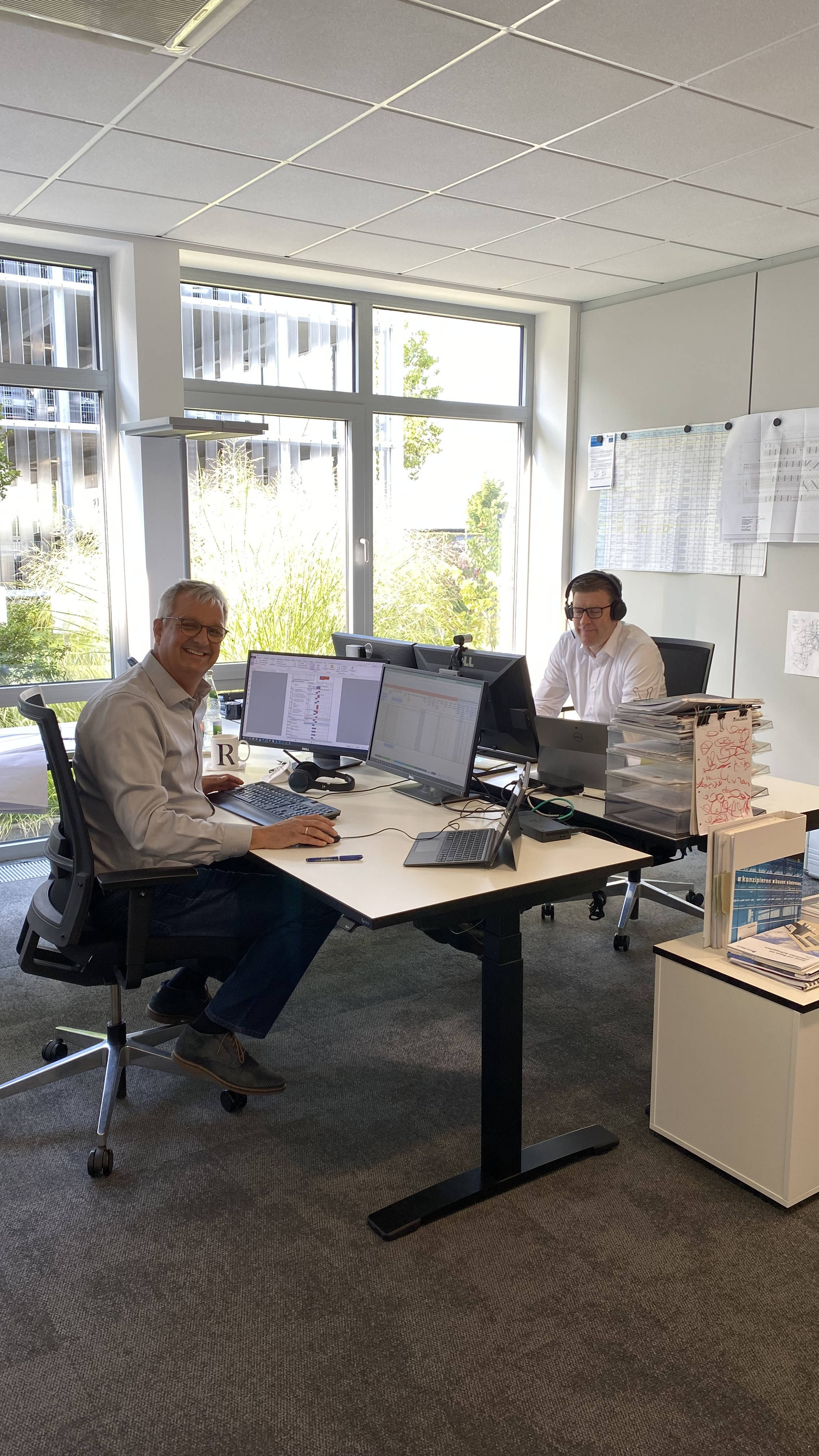 Zwei Männer arbeiten an Computern in einem modernen Büro mit großen Fenstern und technischen Zeichnungen an der Wand.