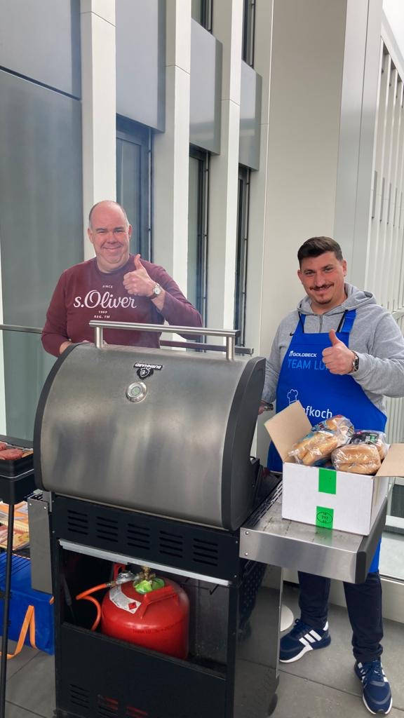Zwei Männer an einem Gasgrill auf einer Terrasse, einer hält Daumen hoch, Brötchen und Burger auf dem Grill.